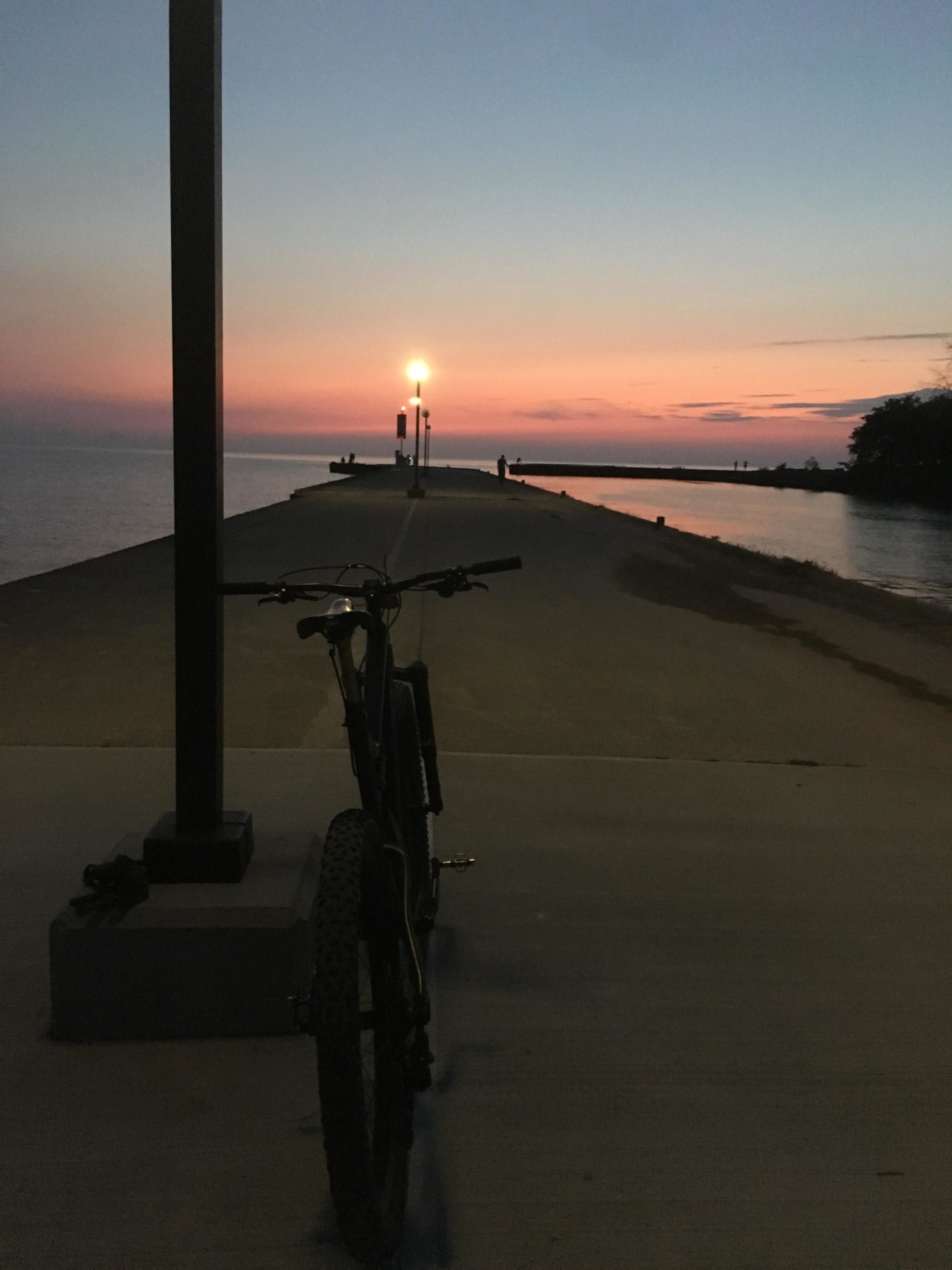 A bicycle parked on a pier during sunset, with a calm body of water and a glowing street lamp in the distance. The sky is filled with hues of pink and blue as twilight approaches. Bayfield river mountain bike trail.