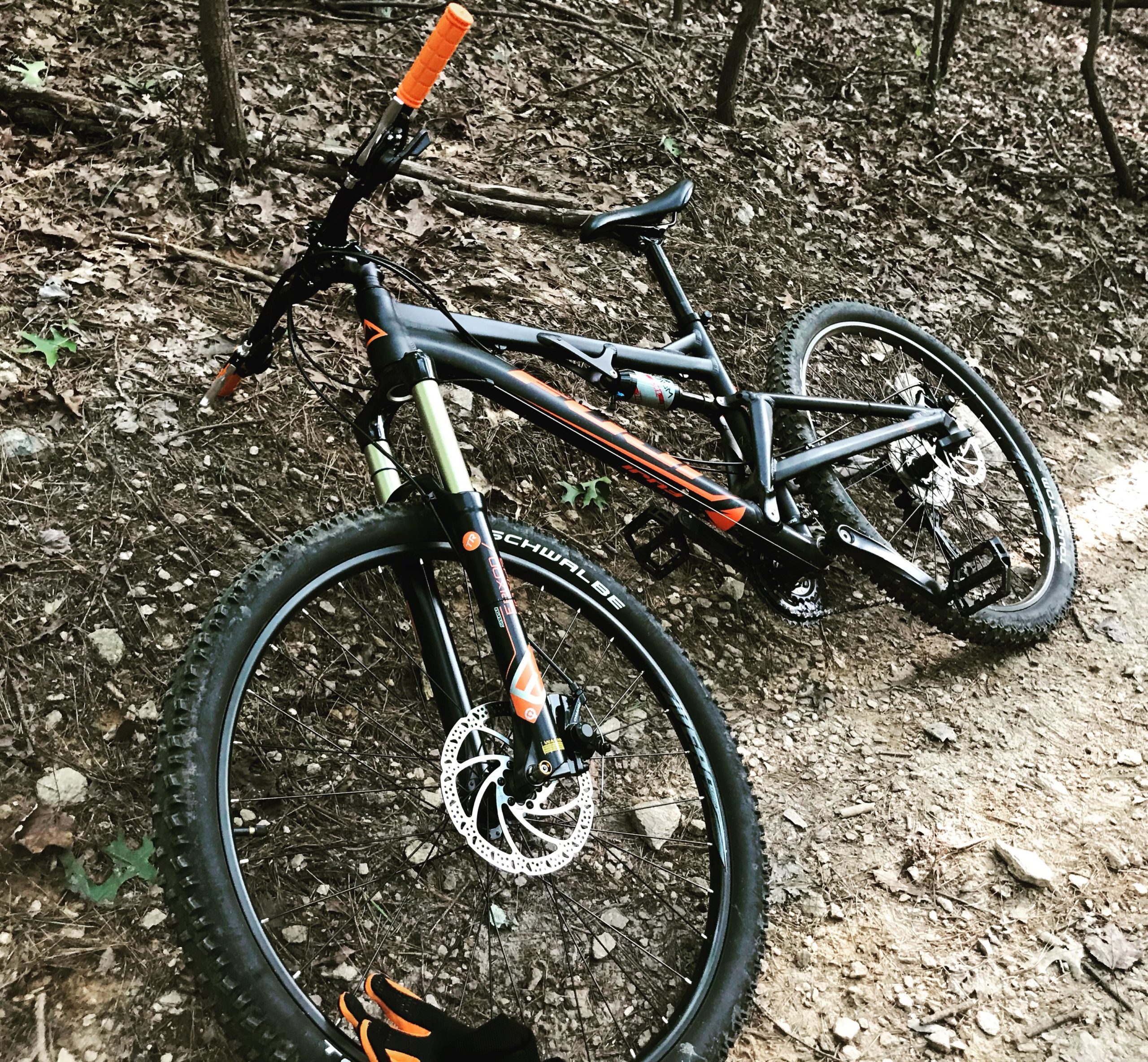 A modern mountain bike with a black and orange frame rests on a dirt trail, surrounded by fallen leaves and greenery. The bike features large tires, a front suspension fork, and disc brakes, with an orange grip on the handlebars. Itusi @ Lake Norman State Park mountain bike trail.