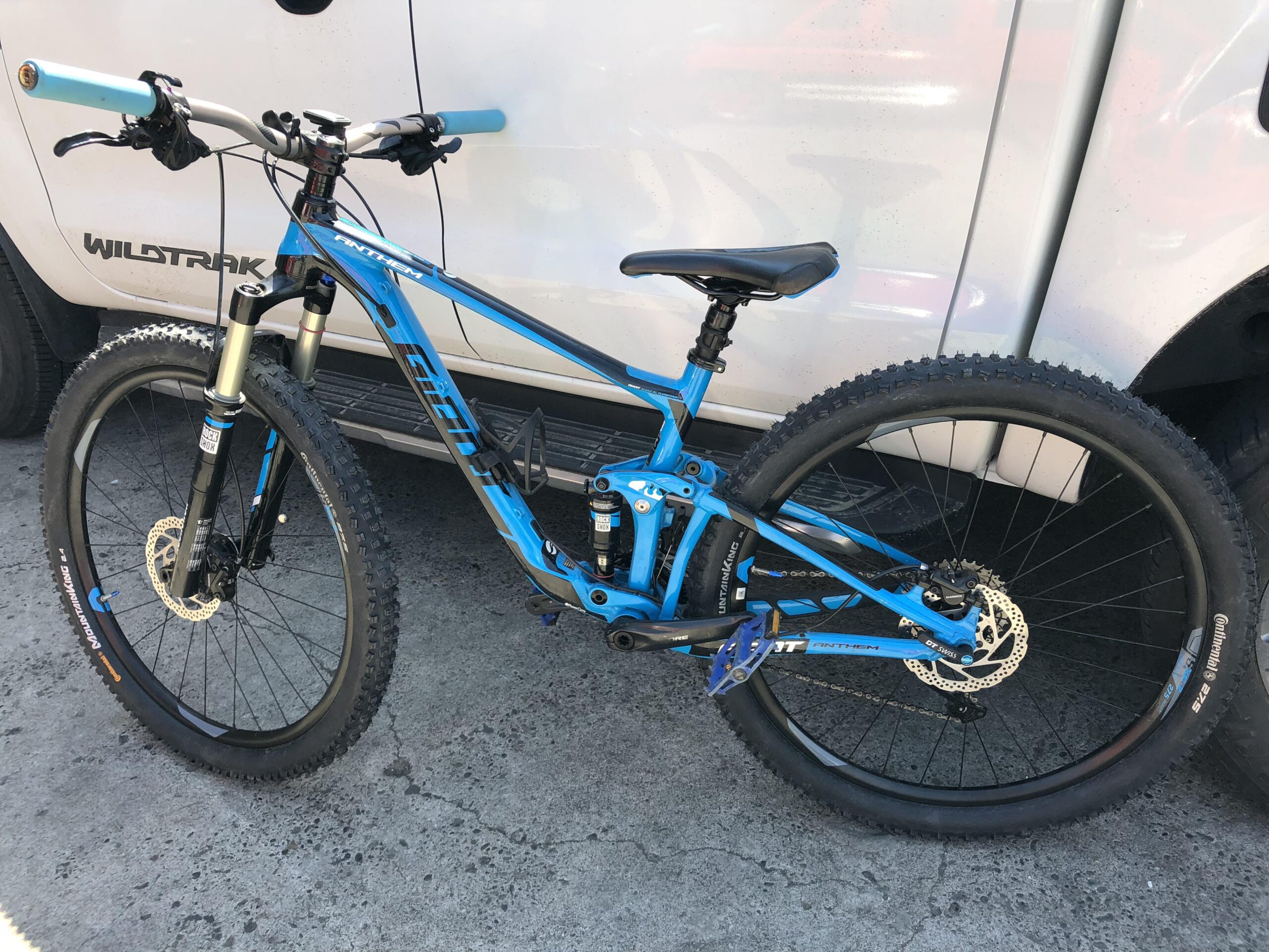 Giant Anthem 27.5 3: A blue mountain bike with a lightweight frame and thick tires, parked next to a white vehicle. The bike features front suspension, a comfortable saddle, and disc brakes. The background shows a concrete surface.