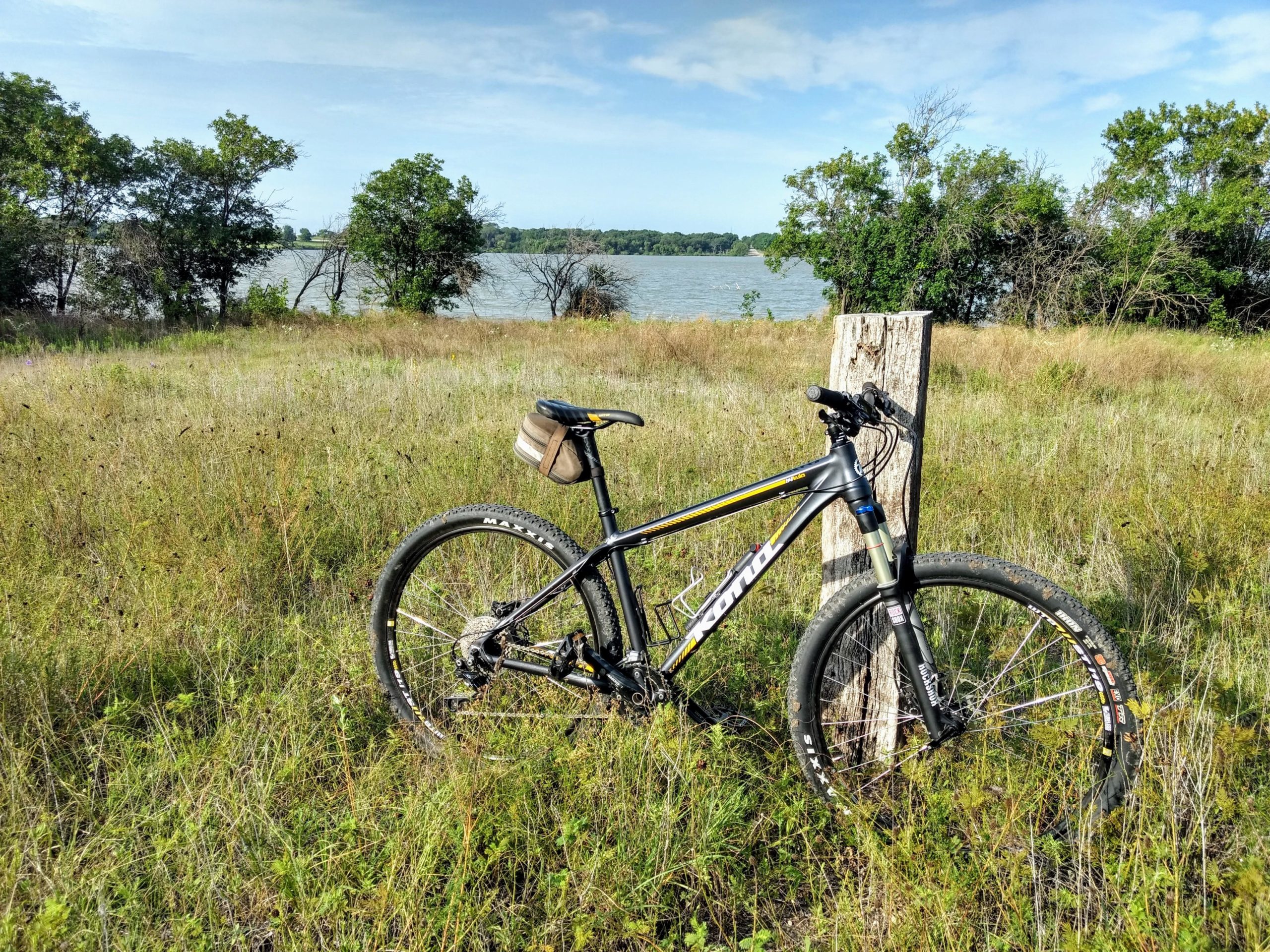 A mountain bike resting against a wooden post in a grassy field, with trees and a lake in the background under a clear blue sky. Mustang Creek Park @ Lake Bardwell mountain bike trail.