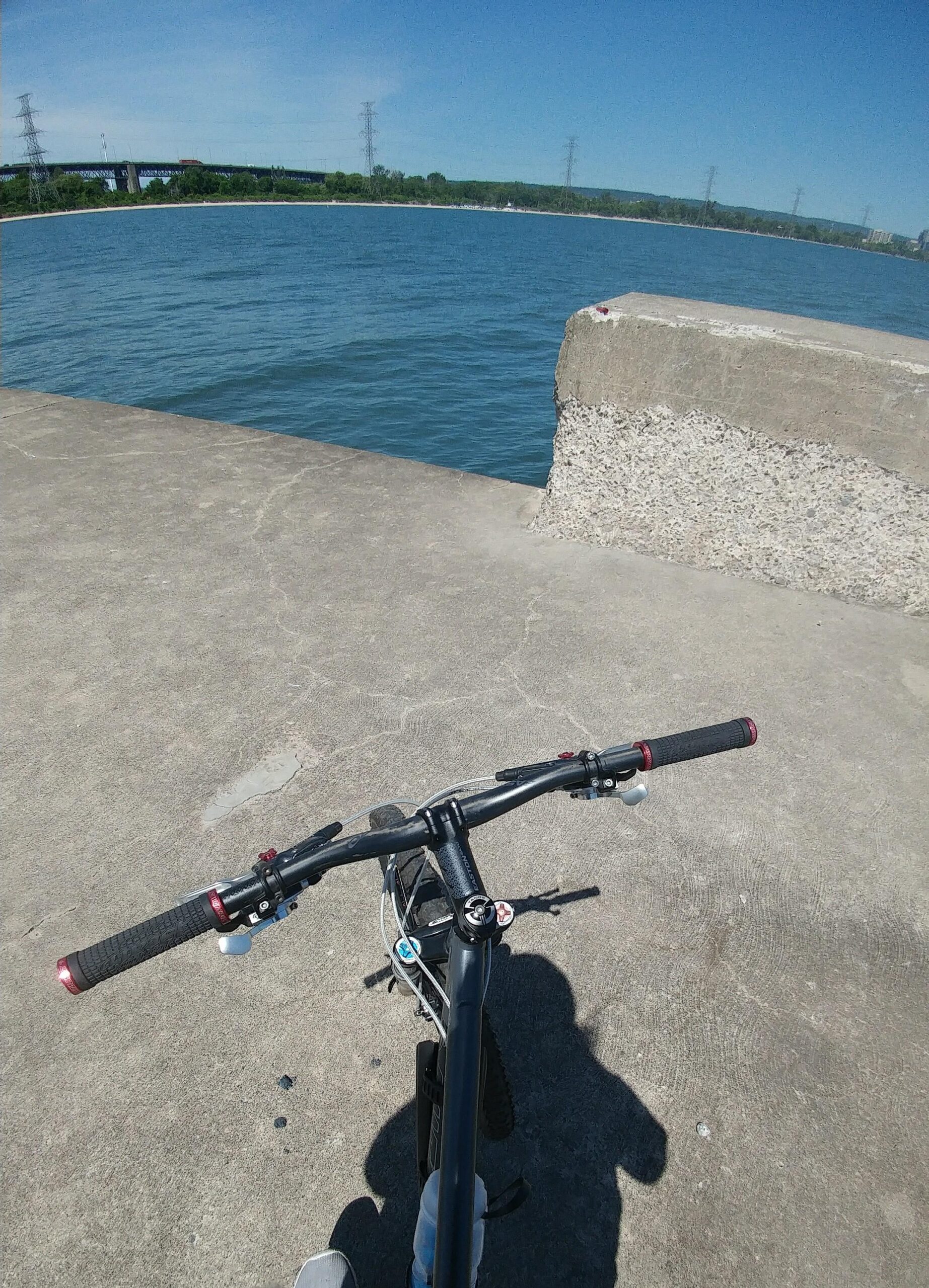 Santa Cruz Blur: A view from the handlebars of a bike overlooking a calm blue lake on a sunny day, with a concrete pathway extending towards the water and power lines in the background.