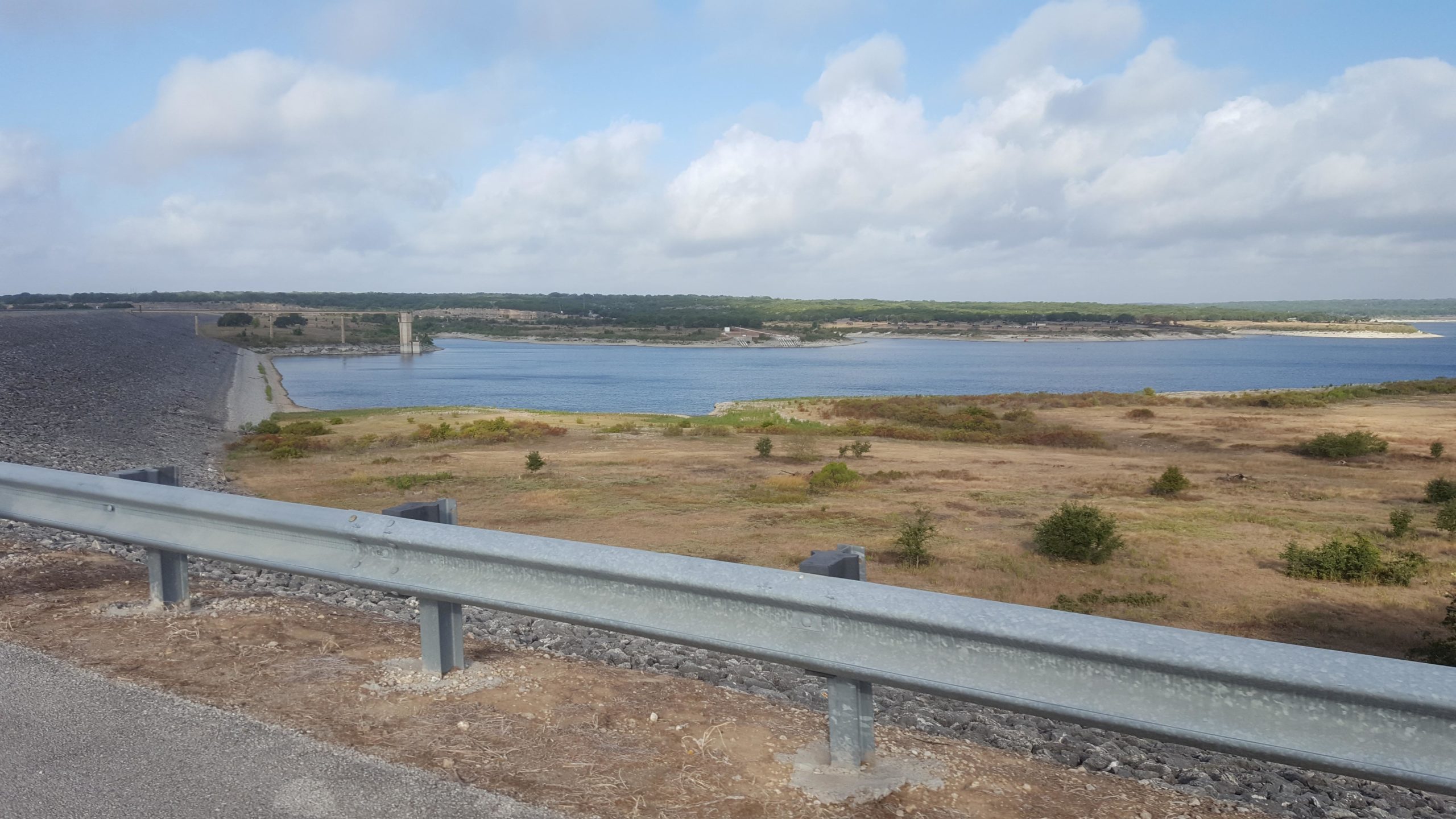 A panoramic view of a tranquil lake bordered by grassy land, with a dam on the left side and a cloudy sky above. A metal guardrail runs along the roadside in the foreground. The landscape features patches of greenery and rolling hills in the background, creating a serene natural setting. Goodwater Trail mountain bike trail.