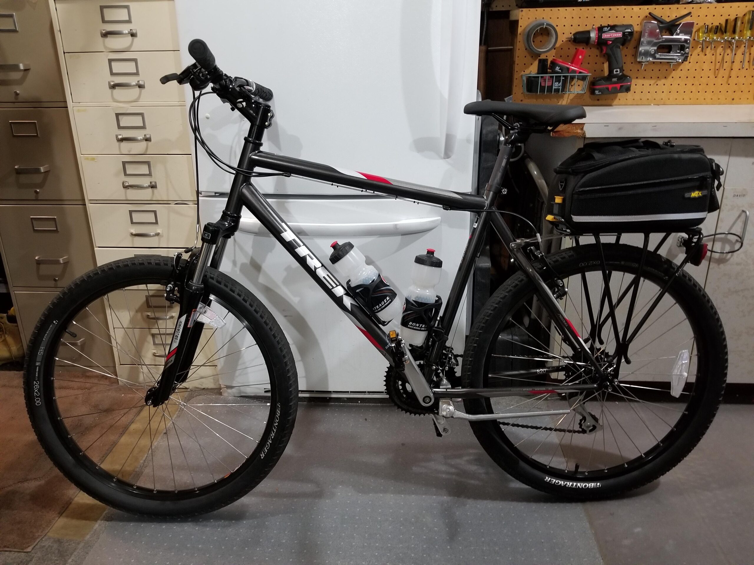 Trek 820: A black Trek mountain bike is leaning against a white refrigerator in a garage. The bike features two water bottle holders, a rear storage bag, and wide tires. In the background, there are metal filing cabinets and a pegboard with various tools, including a drill and tape. The garage floor is gray with sections of different textures.