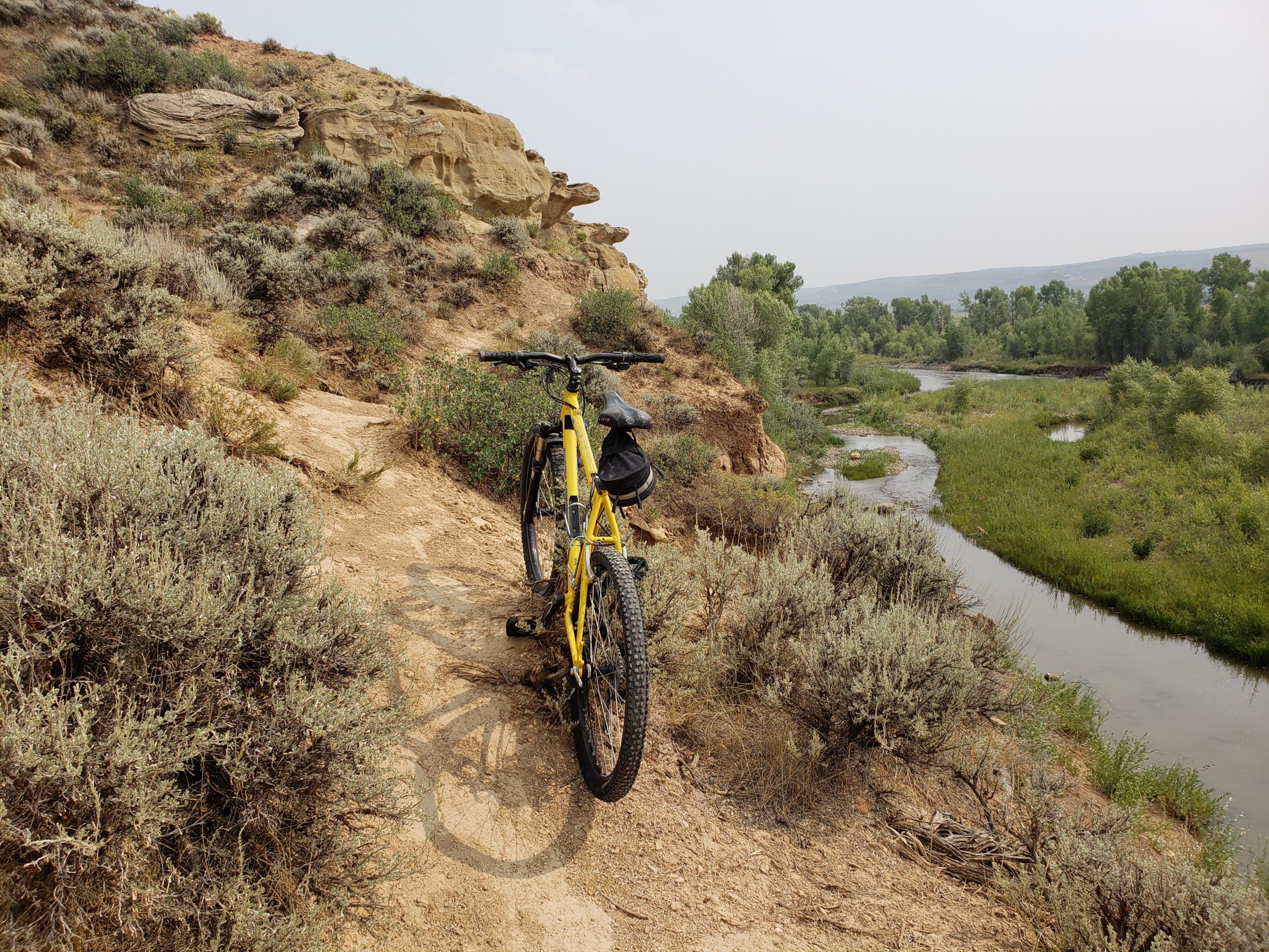 A yellow mountain bike is parked on a dirt trail surrounded by sagebrush and rocky terrain, with a winding river visible in the background under a cloudy sky. Bear River State Park mountain bike trail.