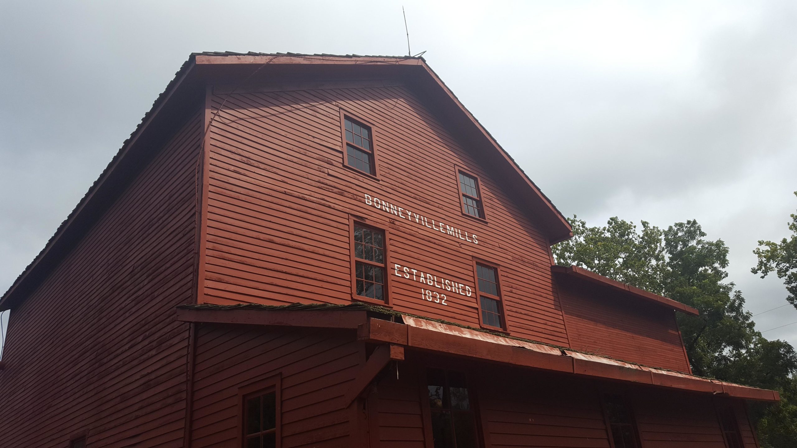 Alt text: A close-up view of the Bonneyville Mills building, featuring red wooden siding and a sign that reads "Bonneyville Mills Established 1832." The image captures the sloped roof and several windows, with a cloudy sky in the background. Bonneyville Mill mountain bike trail.