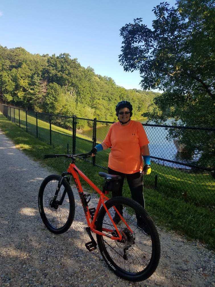 Cannondale Trail 5: A person wearing an orange shirt and gloves stands next to an orange mountain bike on a gravel path by a fenced area, with a body of water visible in the background and a forested hillside beyond. The sky is clear and blue.