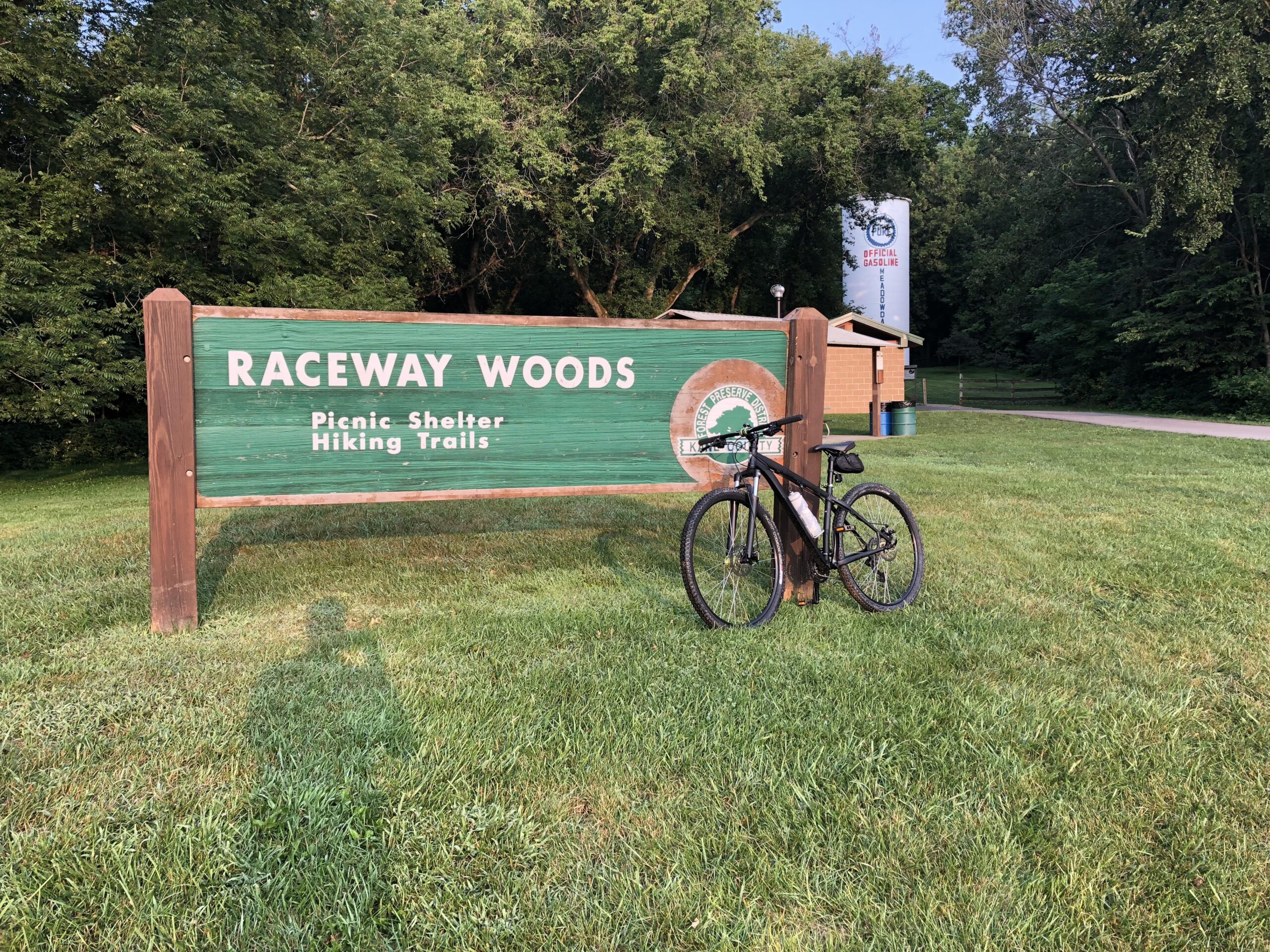 Specialized Rockhopper 29: A wooden sign reading "Raceway Woods" stands in front of a grassy area, with the text "Picnic Shelter" and "Hiking Trails" below it. A black bicycle leans against the sign, surrounded by lush green trees. In the background, there is a building and a path visible, completing a serene outdoor scene.