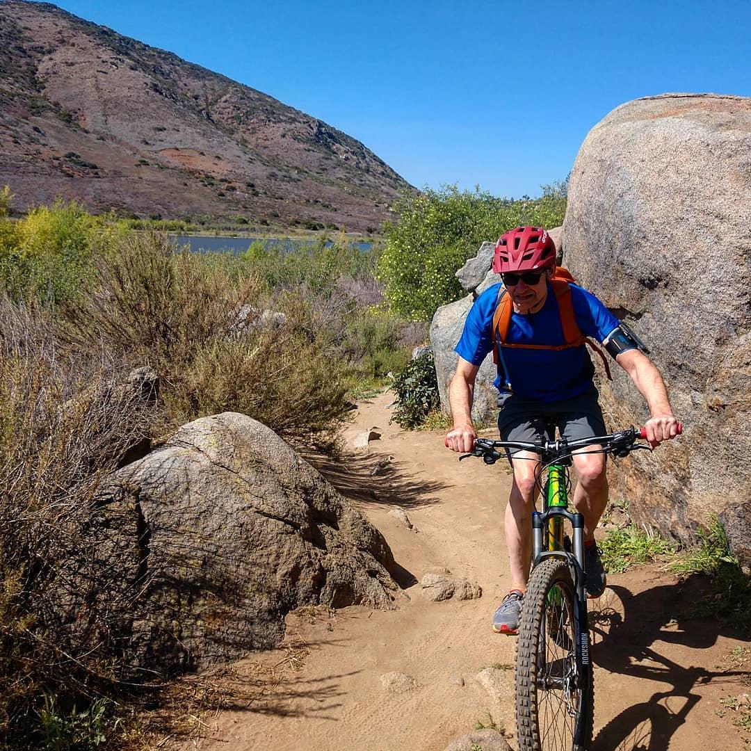 A mountain biker navigating a narrow dirt path between large rocks in a hilly landscape. The rider, wearing a red helmet and blue shirt, appears focused as he pedals through a scenic area with greenery and a body of water in the background under clear blue skies. Lake Hodges mountain bike trail.