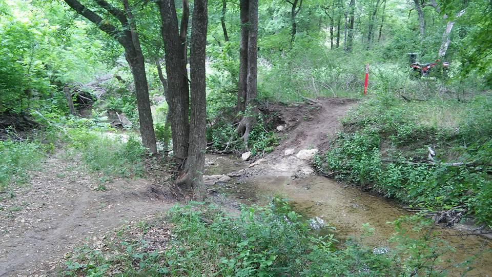 A tranquil forest scene featuring a shallow creek flowing through lush greenery. On the left, a dirt path leads into the woods, while on the right, a small rise leads away from the water. Several trees frame the scene, and a hint of a red marker or post can be seen in the background. The overall atmosphere is serene and natural. Parks of Aledo Trail mountain bike trail.