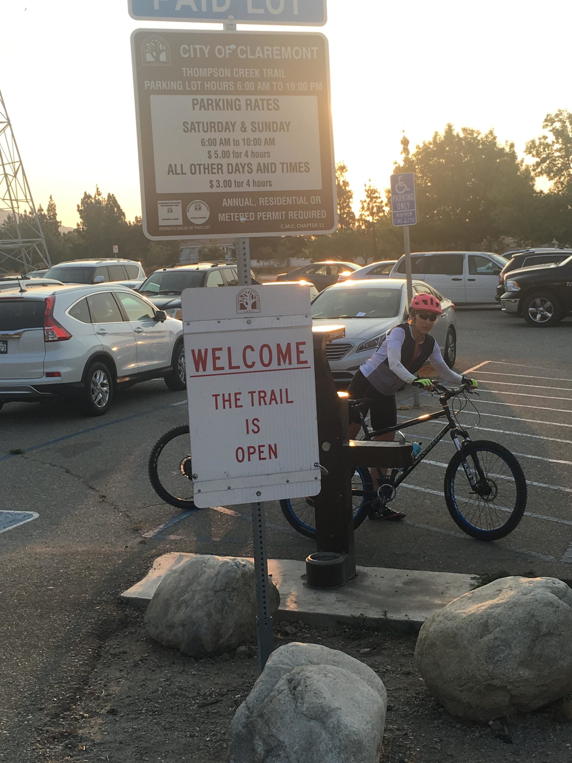 A cyclist in a pink helmet and cycling gear is preparing to ride a mountain bike in a parking lot near a trailhead. In the background, a sign indicates the trail is open, while another sign provides parking regulations and rates for the Thompson Creek Trail in Claremont. The scene is set during a sunset, with parked vehicles and large rocks lining the area. Claremont Wilderness mountain bike trail.