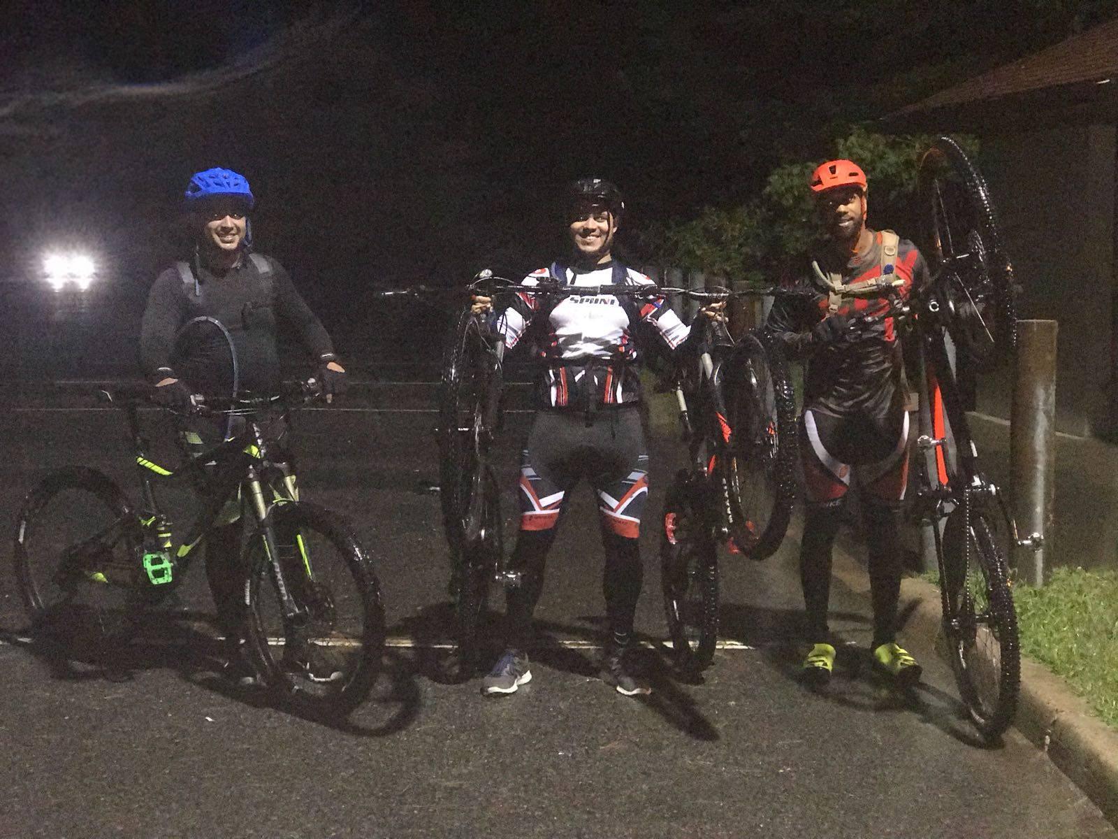 Three cyclists posing together at night, each holding a bike. They are wearing helmets and cycling gear, and there's a dimly lit road in the background with a hint of vehicle headlights. The atmosphere is casual and friendly. Round Valley Recreation Area mountain bike trail.