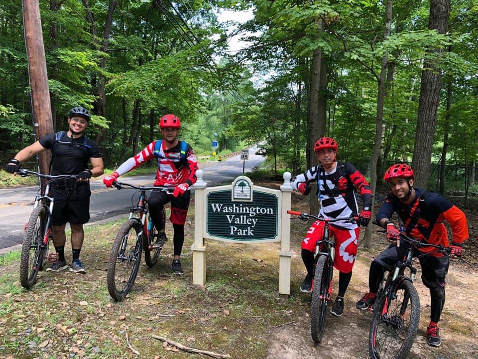 Four mountain bikers pose by a sign for Washington Valley Park, surrounded by lush greenery. Each rider is wearing protective gear and colorful biking apparel, holding their bikes. A road is visible in the background, with trees lining the area. Chimney Rock mountain bike trail.