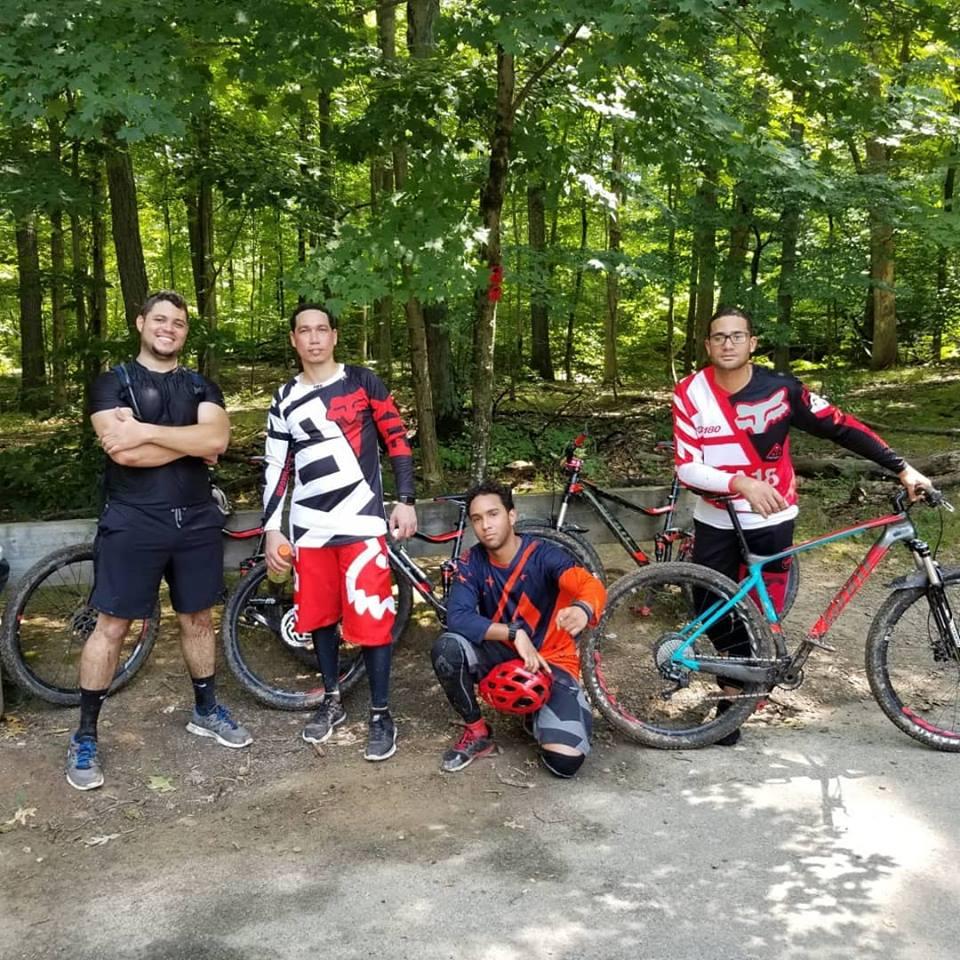 Four mountain bikers posing together in a wooded area. The group is dressed in colorful biking gear, with bikes positioned behind them. They are smiling and appear to be enjoying a break from riding. The background features green trees and a natural setting. Chimney Rock mountain bike trail.
