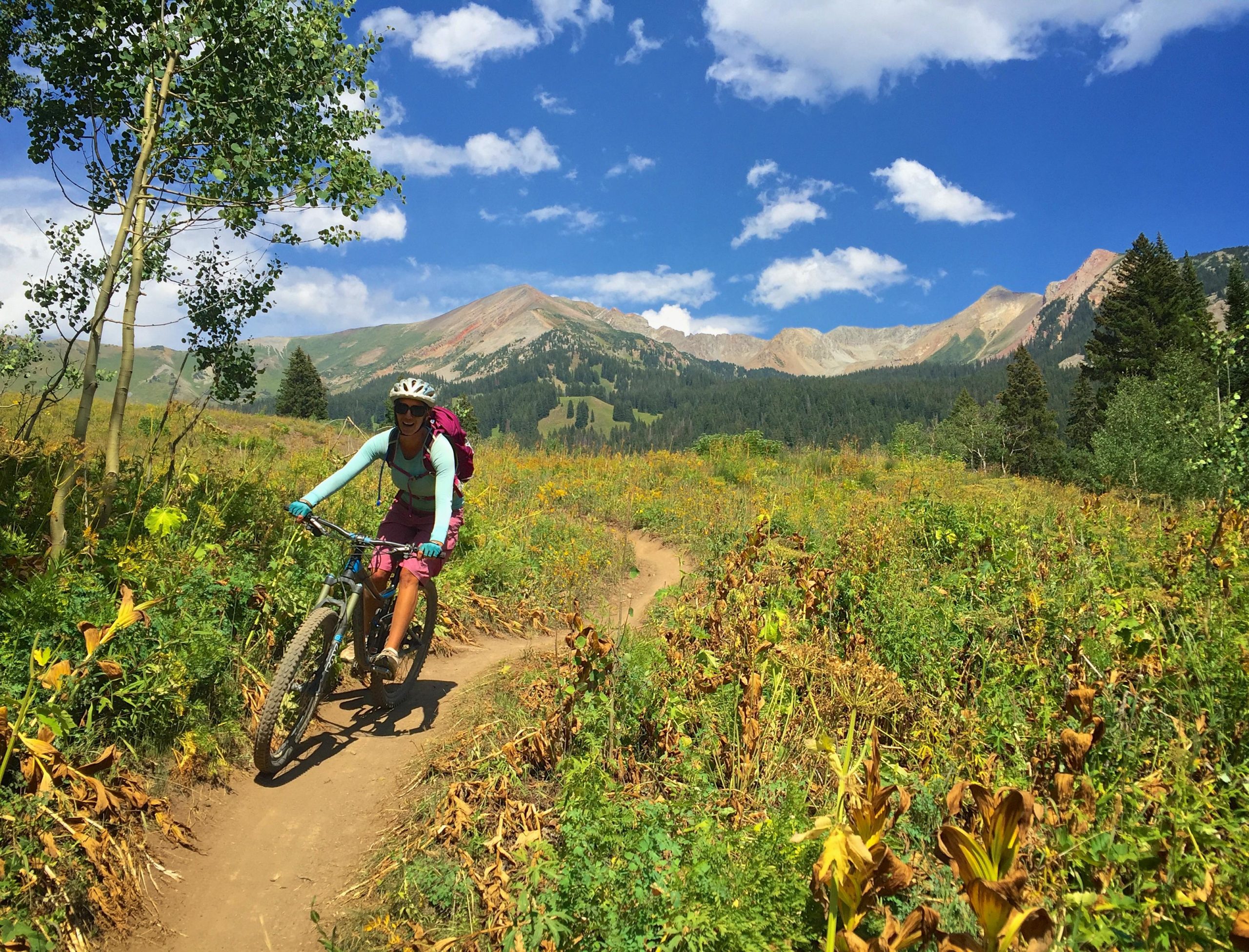 A mountain biker navigating a winding dirt trail through vibrant green foliage, with mountains in the background under a bright blue sky dotted with fluffy clouds. The rider is wearing a helmet and a backpack, smiling as they ride. Trail 401 mountain bike trail.