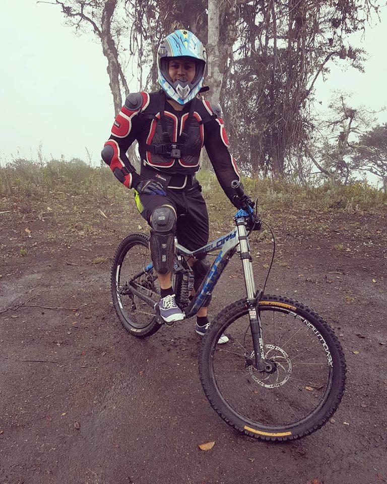 A person wearing a full protective biking outfit, including a helmet, knee pads, and padded chest gear, stands next to a mountain bike on a dirt path. The background features trees and a misty atmosphere, indicating an outdoor environment. Lligua Loop mountain bike trail.