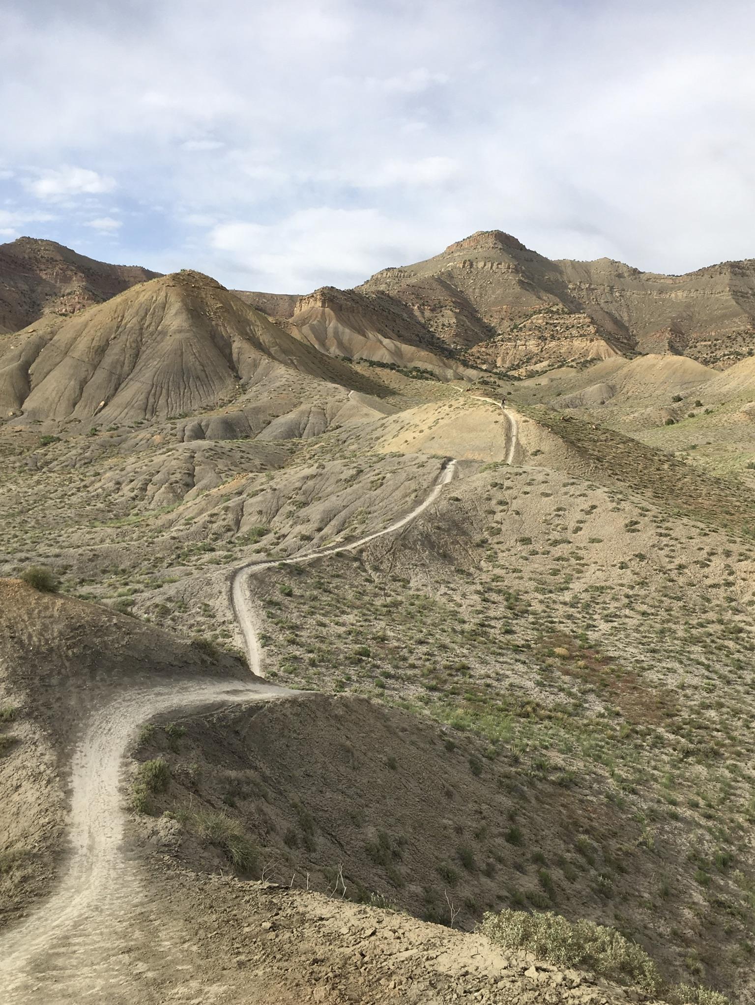 A winding dirt trail leads through arid hills and rugged terrain, under a partly cloudy sky. The landscape features rolling mounds with sparse vegetation, highlighting the natural beauty of a dry mountainous region. Zippety Do Dah mountain bike trail.