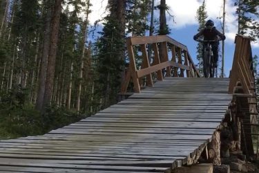 A mountain biker rides up a wooden ramp in a forested area, surrounded by tall trees and a clear blue sky with white clouds. The ramp is constructed from wooden planks and is elevated above the ground, suggesting a challenging biking trail. Trestle Bike Park mountain bike trail.