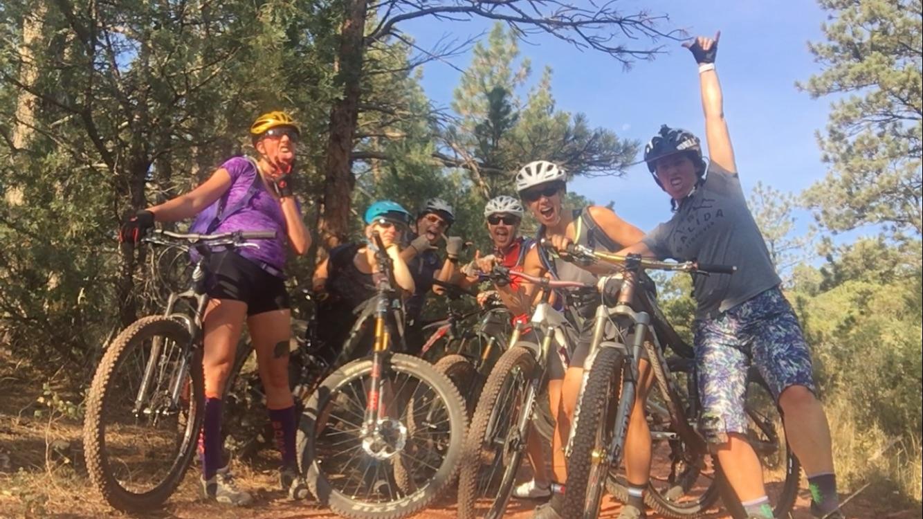 A group of six people poses joyfully on mountain bikes in a forested area, surrounded by trees. Some individuals are making playful gestures, and everyone is wearing helmets and casual biking attire. The scene conveys a sense of camaraderie and adventure in an outdoor setting. Heil Valley Ranch mountain bike trail.