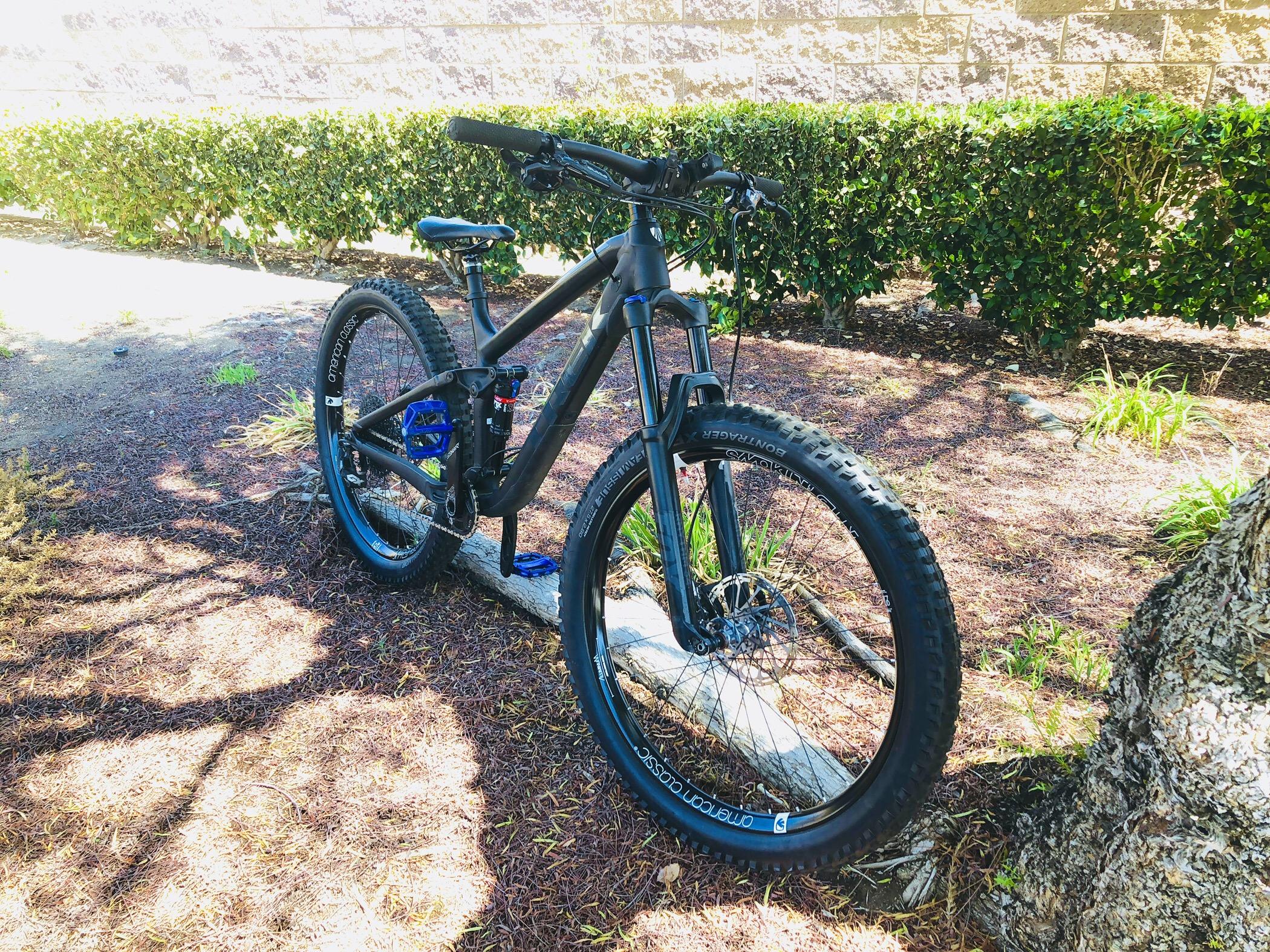 Trek Fuel EX 8: A black mountain bike with blue accents is parked beside a tree in a landscaped area. The bike features wide tires and a sturdy frame, resting on a wooden log amidst foliage and a patch of gravel.
