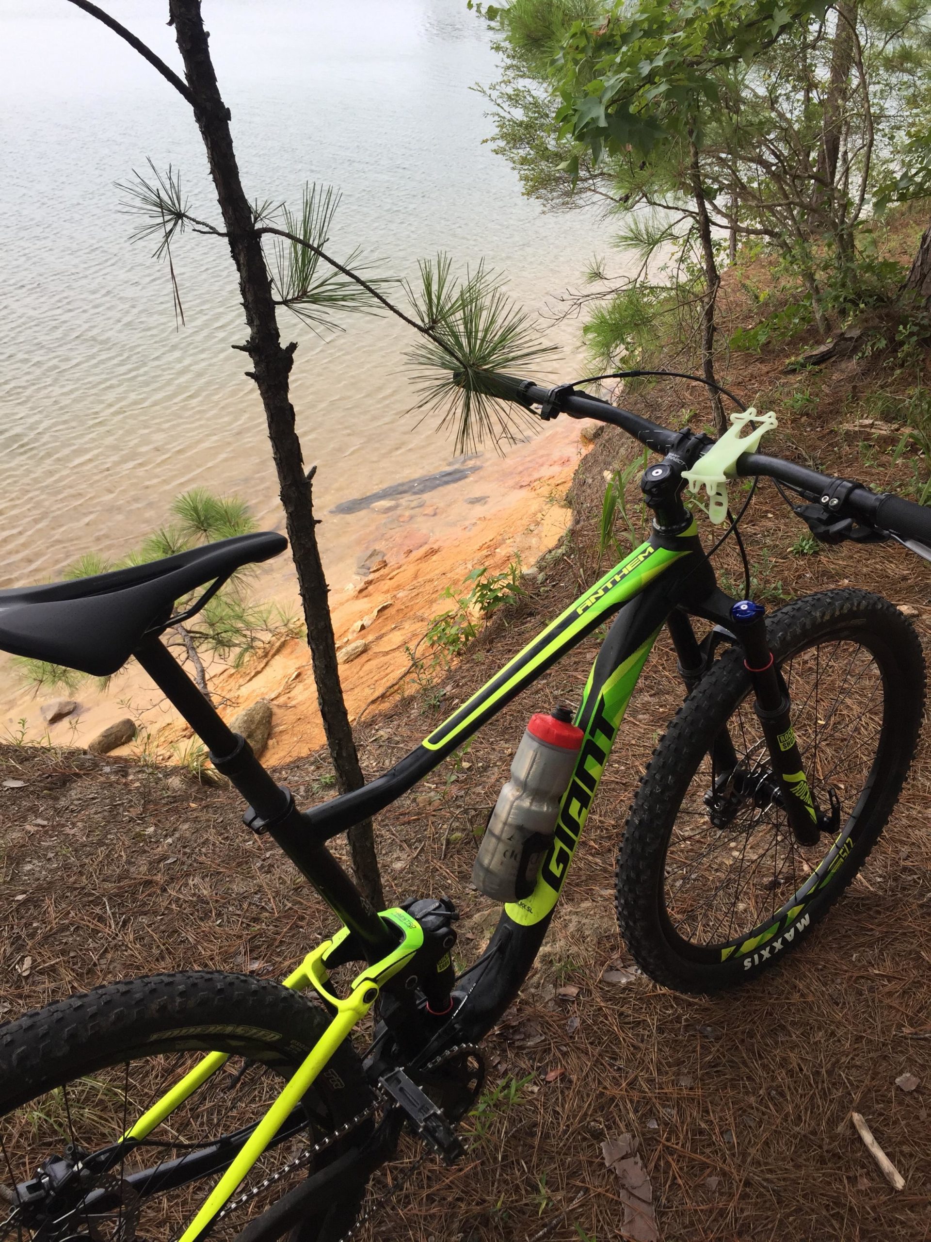Mountain bike positioned near the water's edge, surrounded by trees and natural terrain. The bike features a bright green and black design, with a water bottle attached to the frame. The tranquil lake in the background reflects a calm atmosphere, highlighting the outdoor setting. Bartram Trail / West Dam / Wildwood Park mountain bike trail.