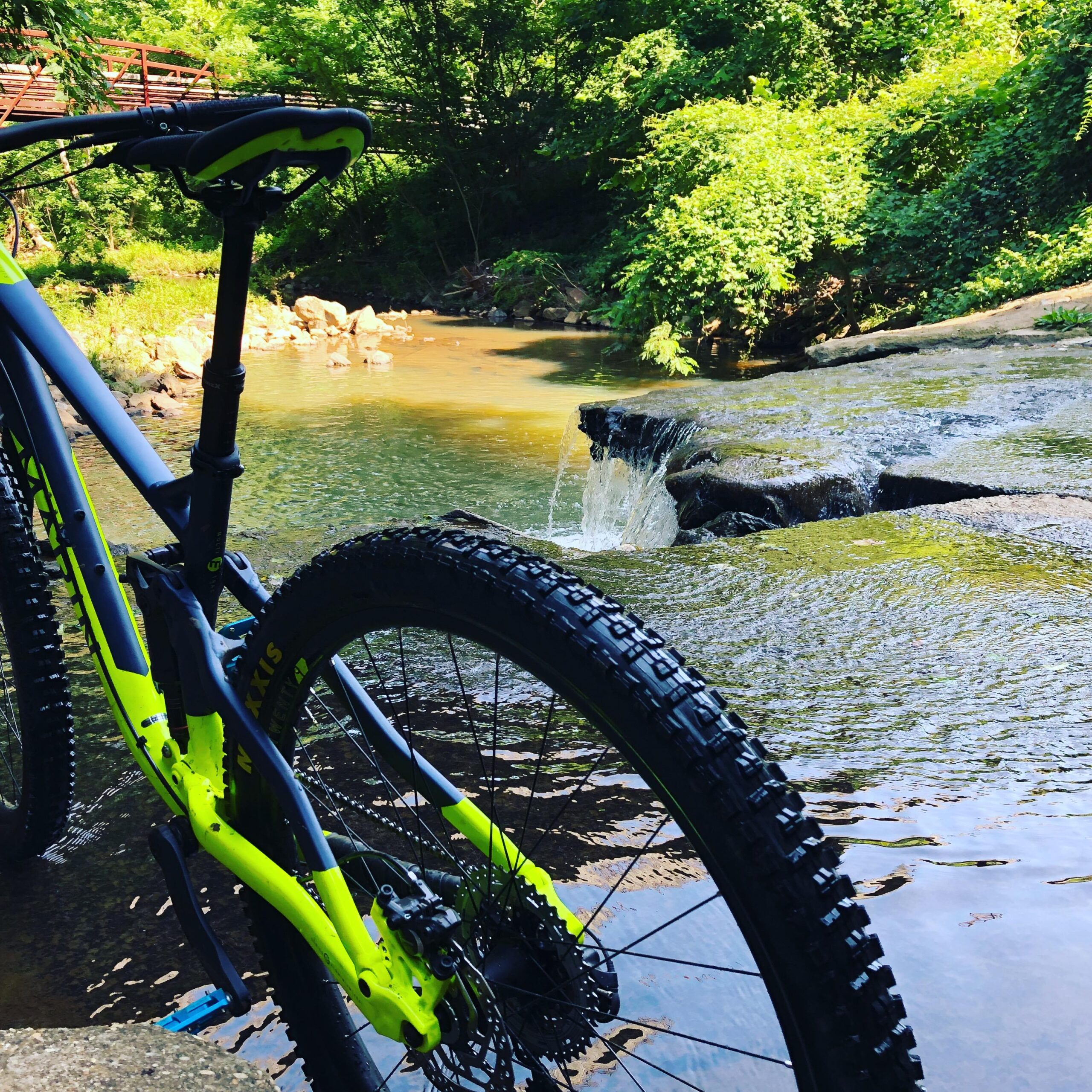 Marin Rift Zone 29er: A close-up view of a mountain bike with a brightly colored frame parked near a creek. The background features a gently flowing stream with small rocks and a cascade of water, surrounded by lush green foliage and trees. A wooden bridge is visible in the distance, adding to the natural scenery.
