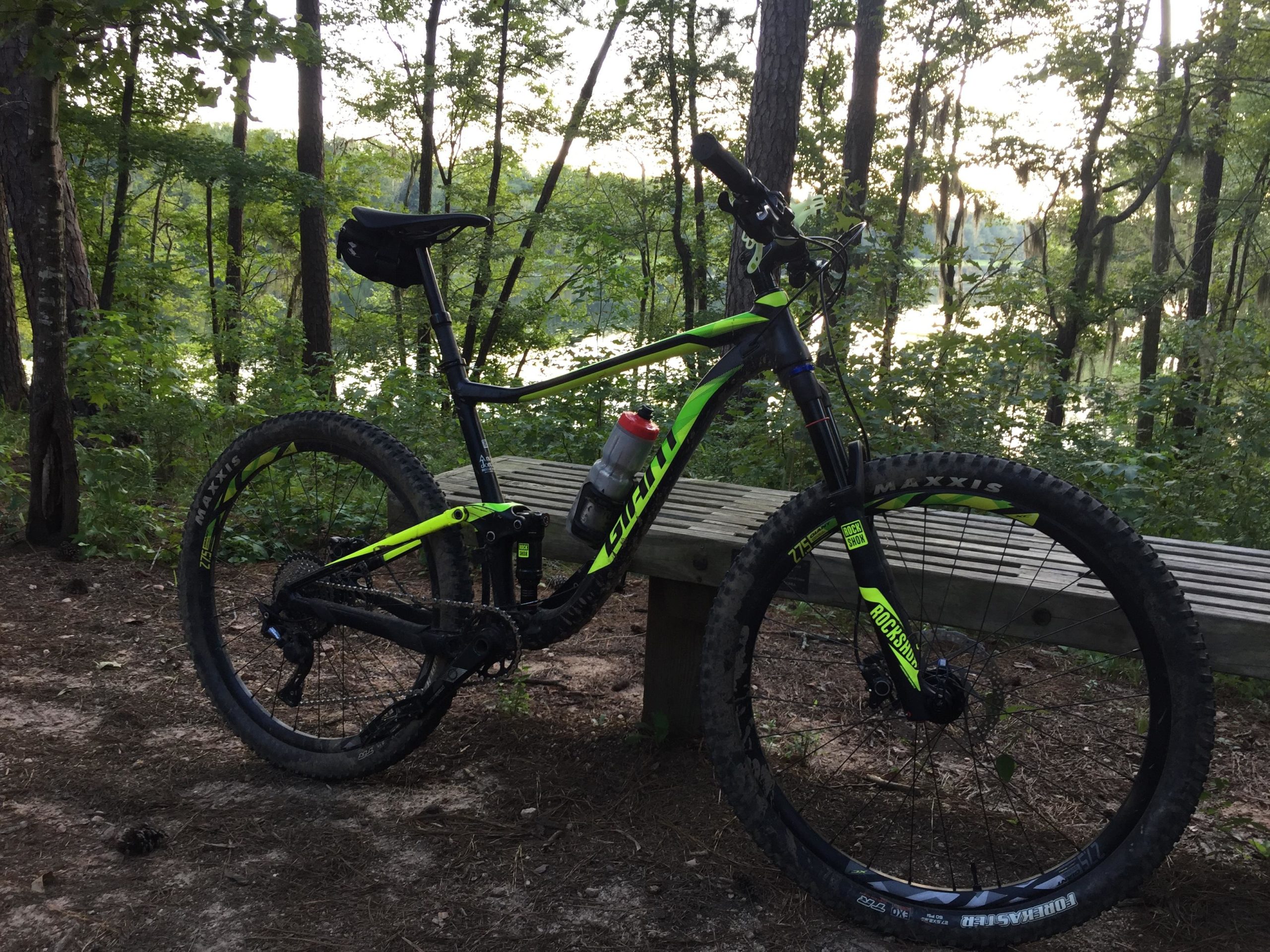 A mountain bike with a black and bright green frame is parked next to a wooden bench in a wooded area. In the background, trees frame a serene view of a lake, suggesting a tranquil outdoor setting for biking. A water bottle is attached to the bike, emphasizing its readiness for an adventurous ride. Forks Area Trail System (FATS) mountain bike trail.