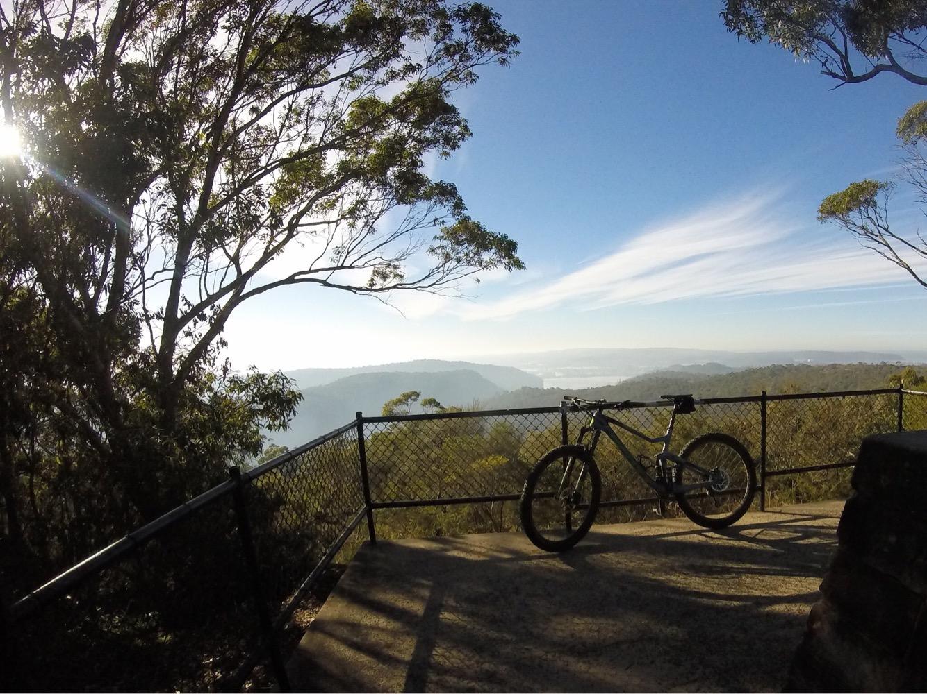 A mountain bike rests on a viewing platform surrounded by trees, overlooking a picturesque valley with rolling hills, under a clear blue sky. The sun shines through the foliage, creating a bright and inviting atmosphere. Brisbane Water National Park mountain bike trail.