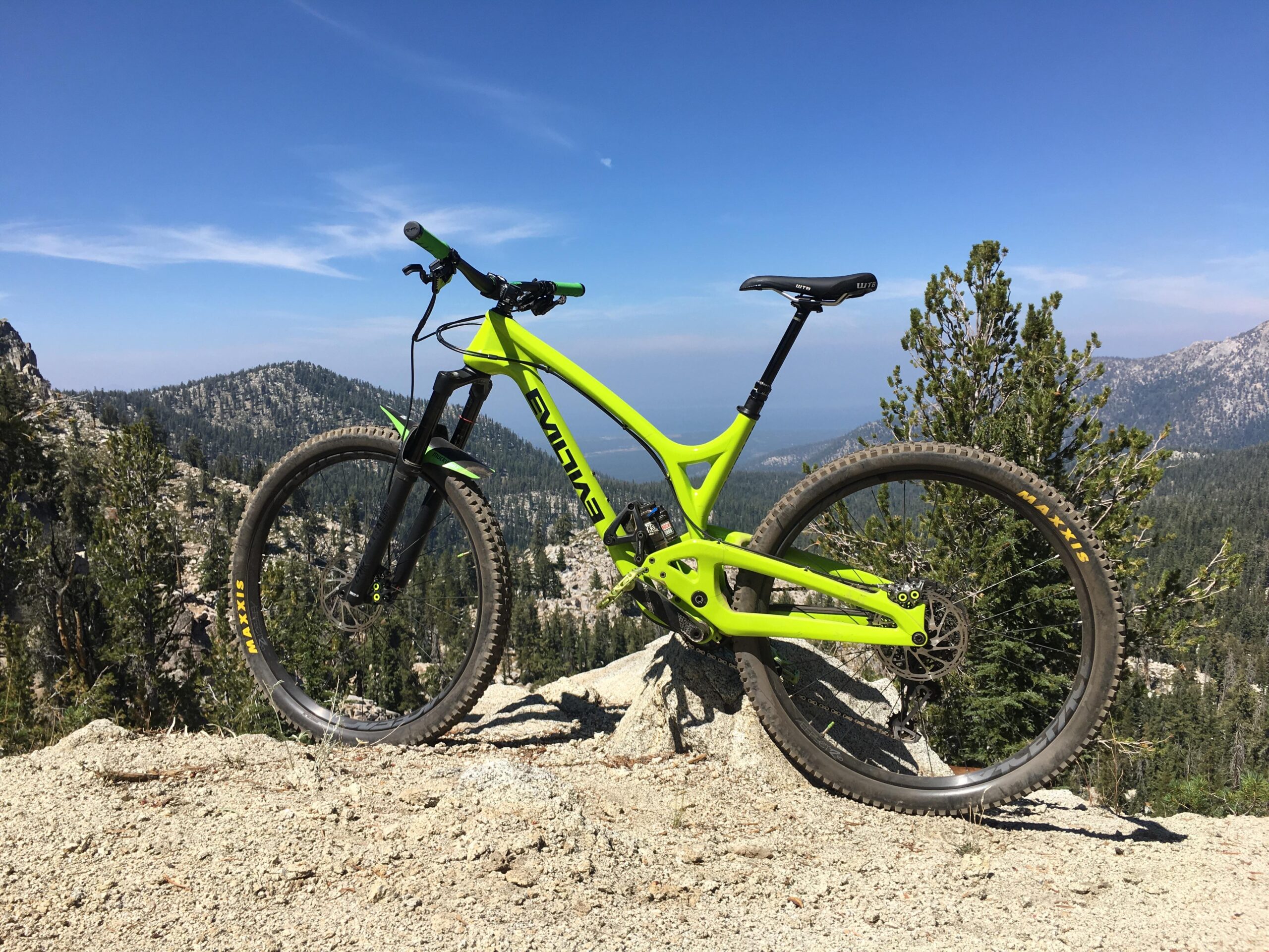 Evil Wreckoning: A bright green mountain bike positioned on rocky terrain with a scenic mountainous landscape in the background, under a clear blue sky.