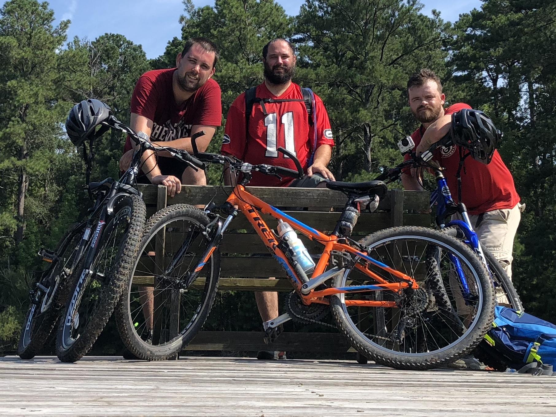 Three men stand on a wooden platform surrounded by trees, posing with their mountain bikes. Two bikes are on the ground beside them—one orange and one blue. The men are wearing casual attire, with one man holding a helmet and another wearing a small camera attached to his bike. The sky is clear and sunny, indicating a nice day for outdoor activities. Children's Home / Pig Trail mountain bike trail.