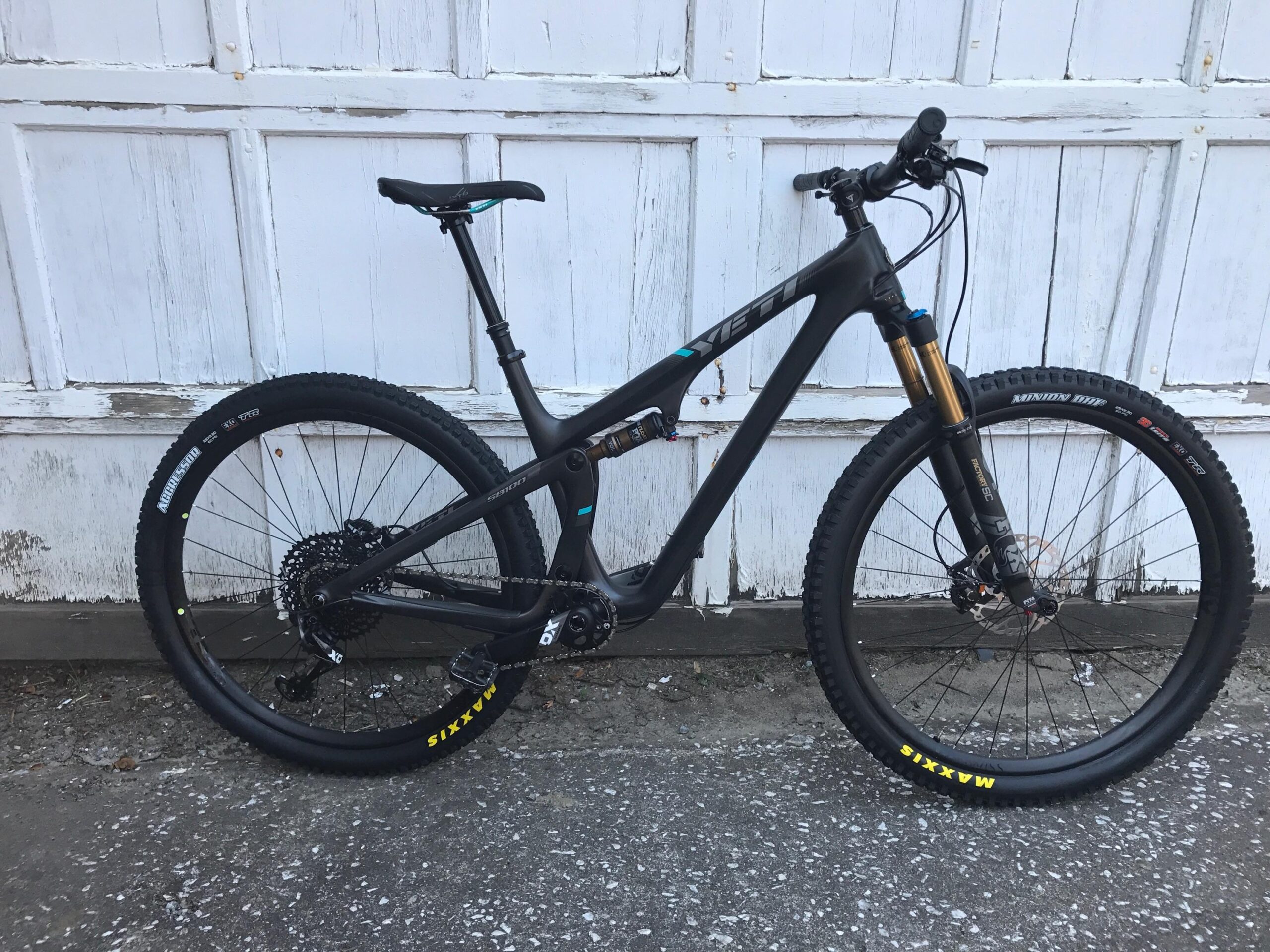 Yeti SB100: A black mountain bike with a sleek design is positioned against a weathered white wooden garage door. The bike features notable components such as thick tires with Maxxis branding, a prominent front fork, and a clean gear setup. The overall aesthetic highlights its performance-oriented build, ideal for rugged terrain.