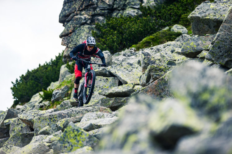 A mountain biker navigating a rocky trail in a mountainous landscape, surrounded by boulders and green shrubbery. The rider is wearing a helmet and protective gear, showcasing a dynamic posture as they maneuver over the challenging terrain.