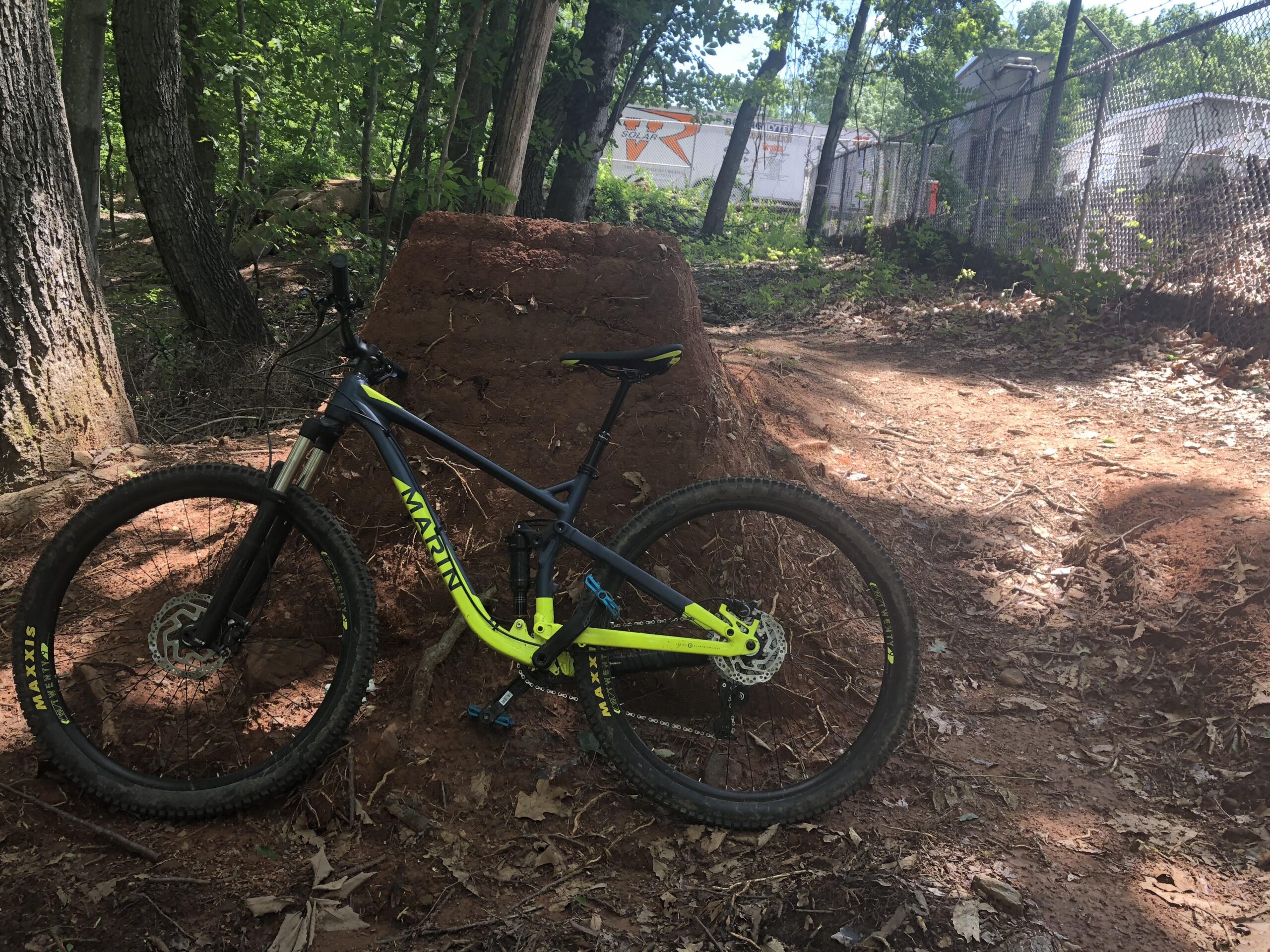 Marin Rift Zone 29er: A blue and yellow mountain bike leaning against a dirt ramp in a wooded area, with trees in the background and a path visible. Sunlight filters through the leaves, creating a dappled light effect on the ground.