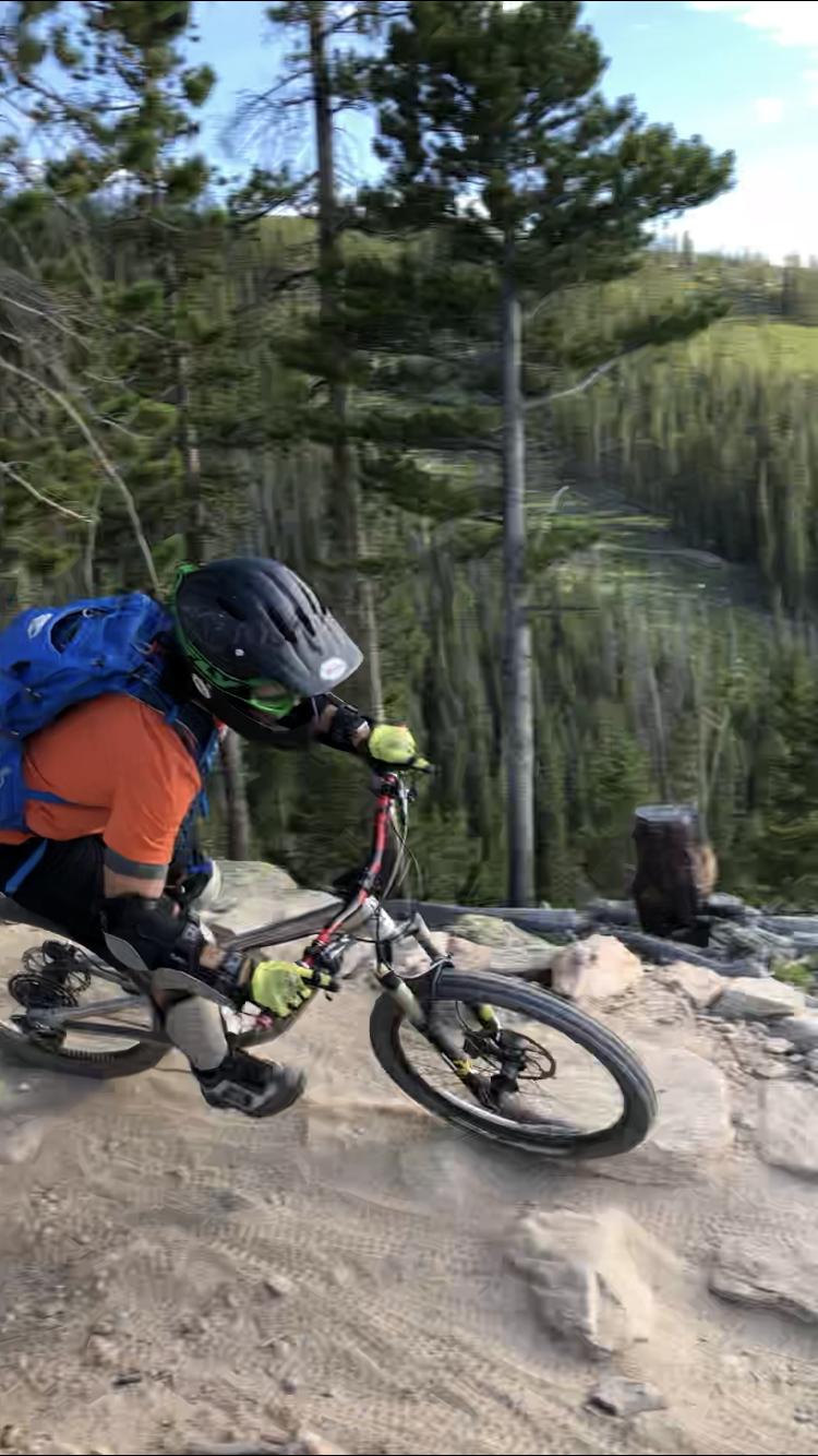 A mountain biker navigates a rocky downhill trail surrounded by tall trees and a scenic forest backdrop, wearing protective gear and a blue backpack. The bike kicks up dust as the rider leans into the turn, showcasing a dynamic and adventurous outdoor experience. Trestle Bike Park mountain bike trail.