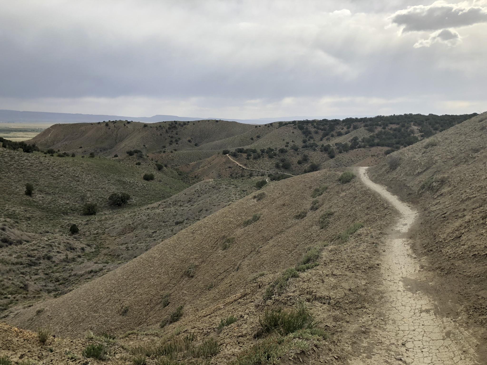 A scenic view of undulating hills and a winding dirt path, under a cloudy sky. The landscape features sparse vegetation and patches of green, with distant hills visible in the background. The terrain appears dry and rugged, indicative of a natural outdoor environment. Zippety Do Dah mountain bike trail.