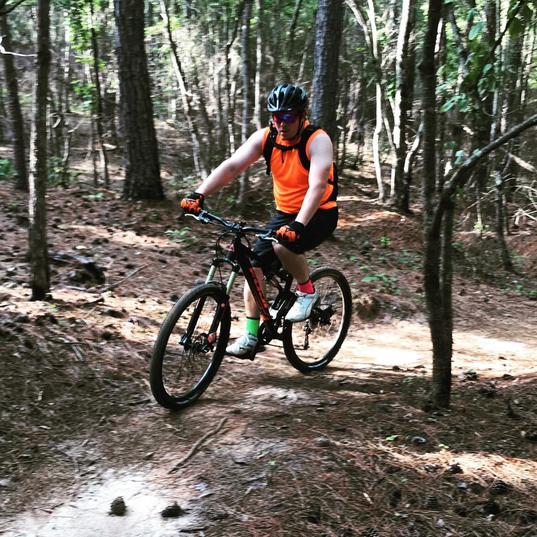 A person riding a mountain bike along a narrow trail in a forested area, wearing an orange sleeveless shirt, black shorts, and a helmet. The scene features tall trees and a carpet of pine needles on the ground, indicating a sunny day. Itusi @ Lake Norman State Park mountain bike trail.