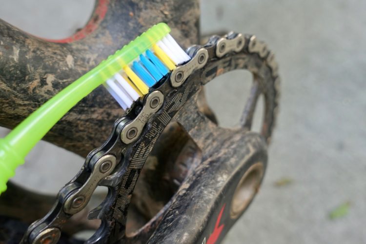 A close-up image of a dirty bicycle chain being cleaned with a colorful toothbrush, highlighting the chain links and the bristles of the toothbrush.