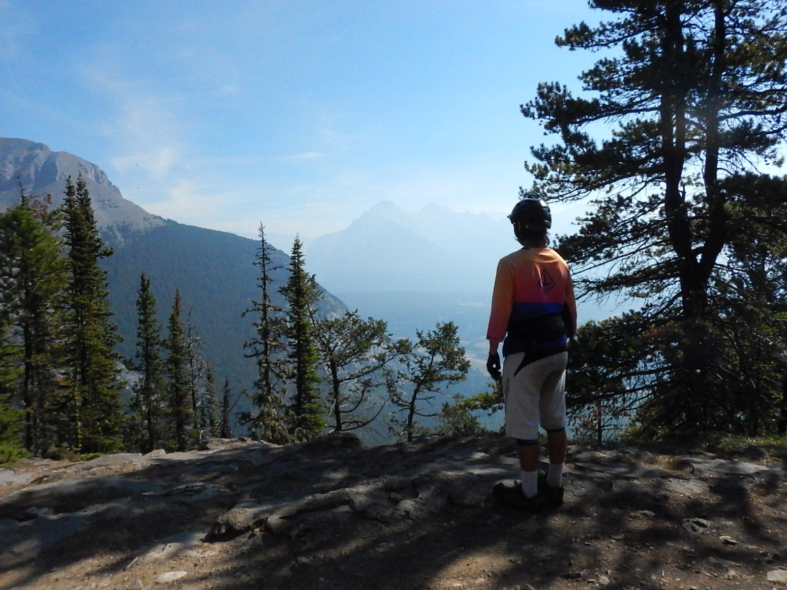 A person standing on a rocky ledge overlooking a mountainous landscape, surrounded by tall trees. The individual is wearing a colorful long-sleeve shirt, shorts, and a helmet, gazing at the distant mountains under a clear blue sky. Stoney Squaw Upper & Lower mountain bike trail.