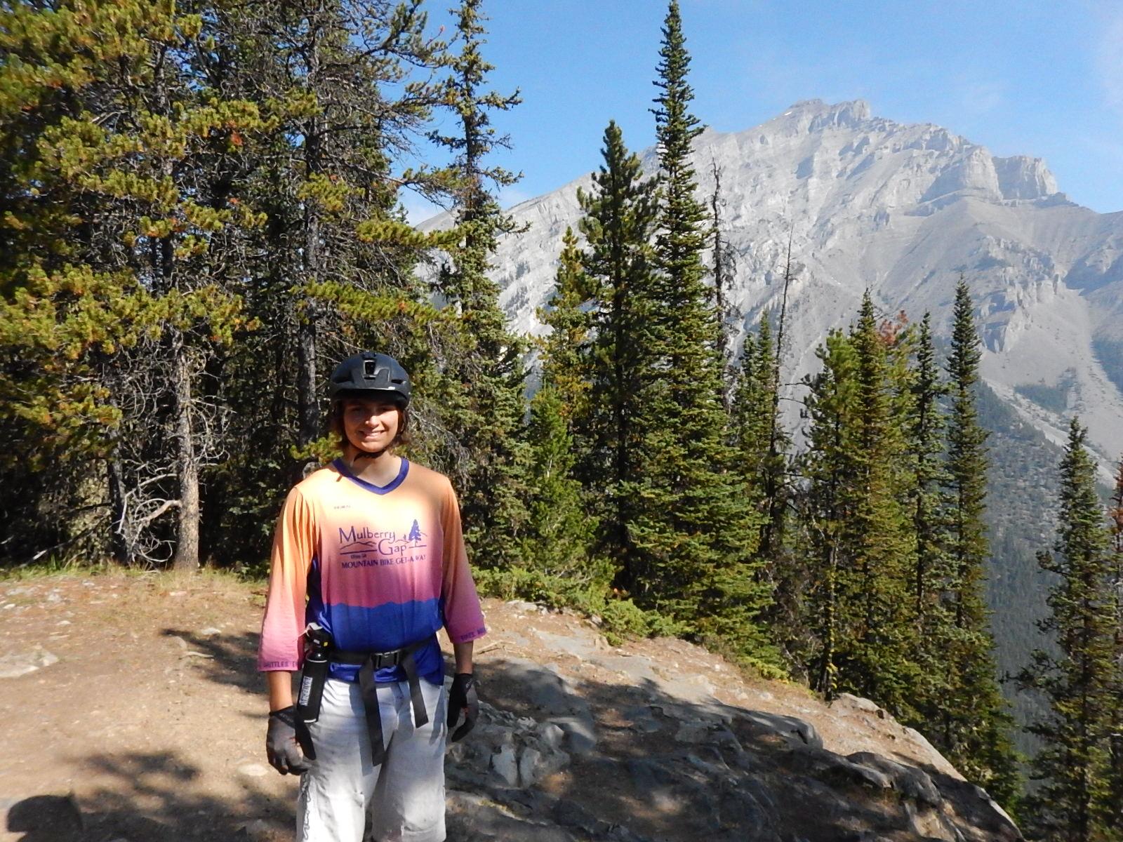 A smiling person wearing a helmet and a colorful biking jersey stands on a rocky path surrounded by tall trees, with a mountain in the background under a clear sky. Stoney Squaw Upper & Lower mountain bike trail.