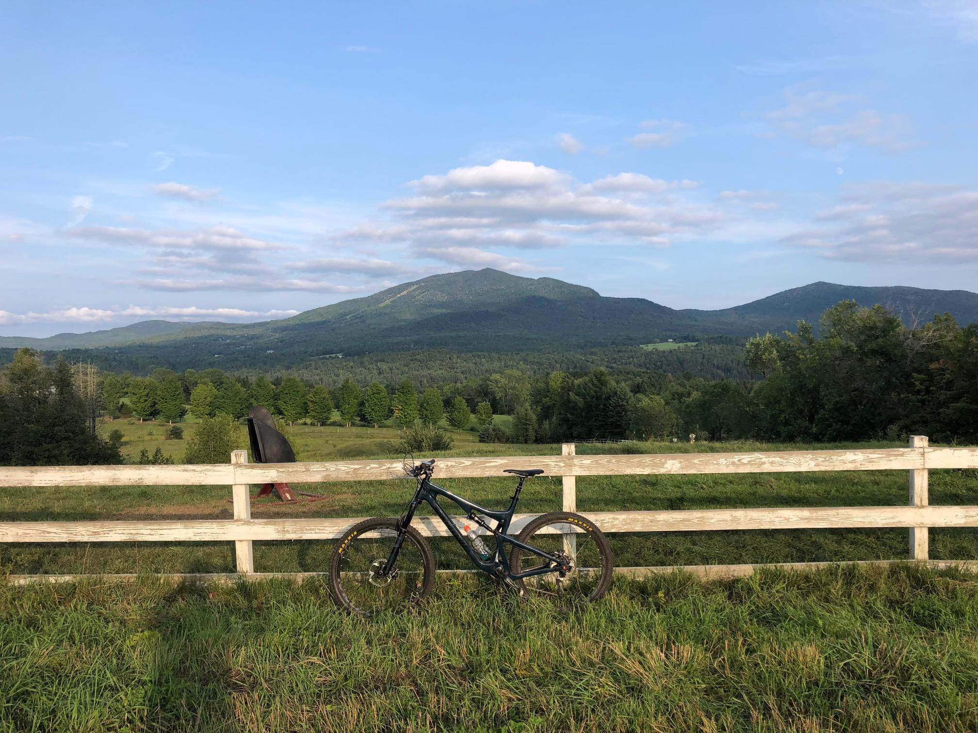 A scenic view featuring a mountain landscape under a blue sky with scattered clouds. In the foreground, a black mountain bike leans against a white wooden fence, with lush green grass and trees in the surrounding area. The mountains rise majestically in the background, showcasing the beauty of nature. Kingdom Trails mountain bike trail.