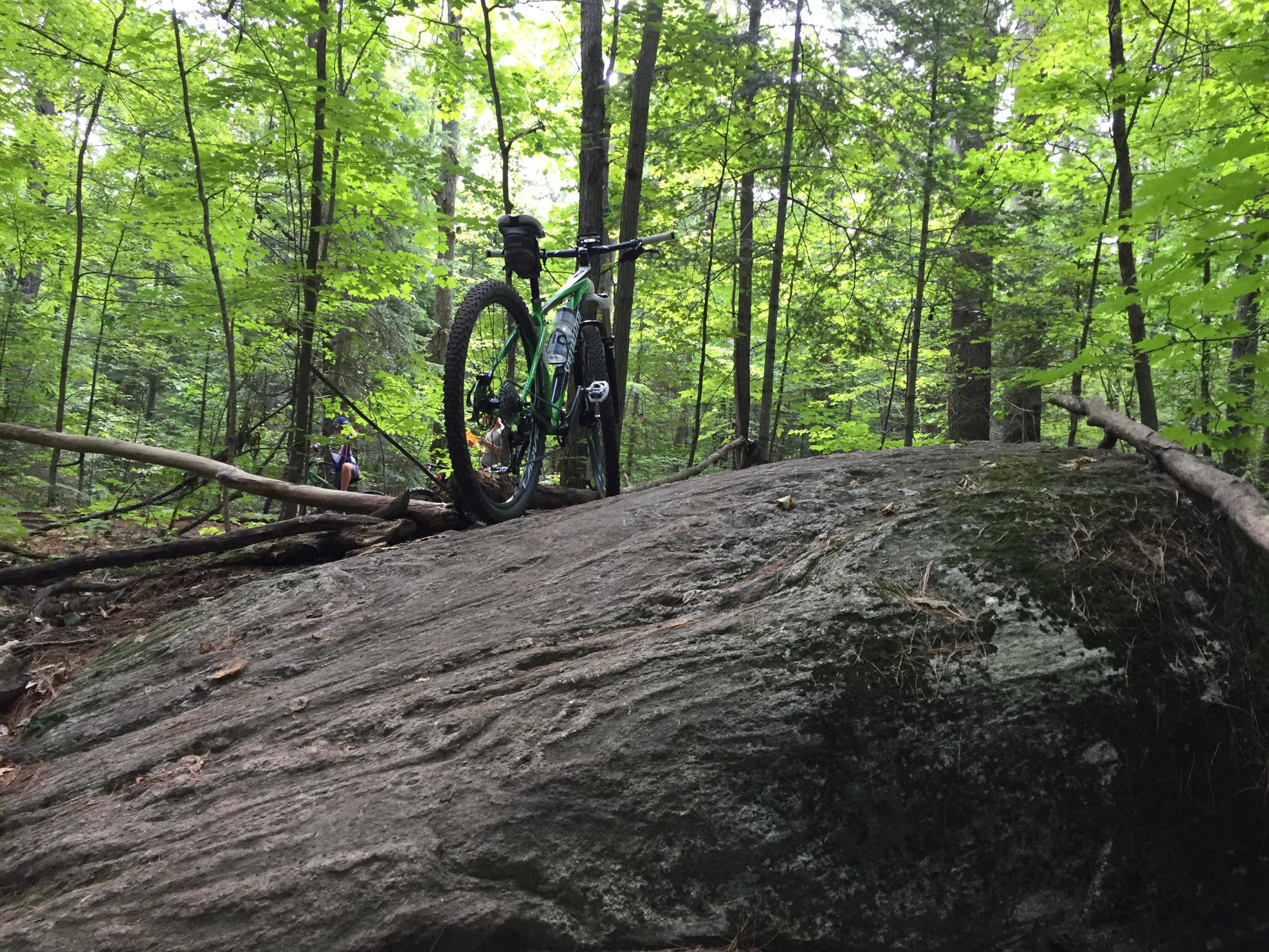 A mountain bike resting on a large rock in a dense forest, surrounded by green trees and foliage. In the background, a person is partially visible among the trees. The scene conveys a sense of adventure and nature. Buckwallow mountain bike trail.