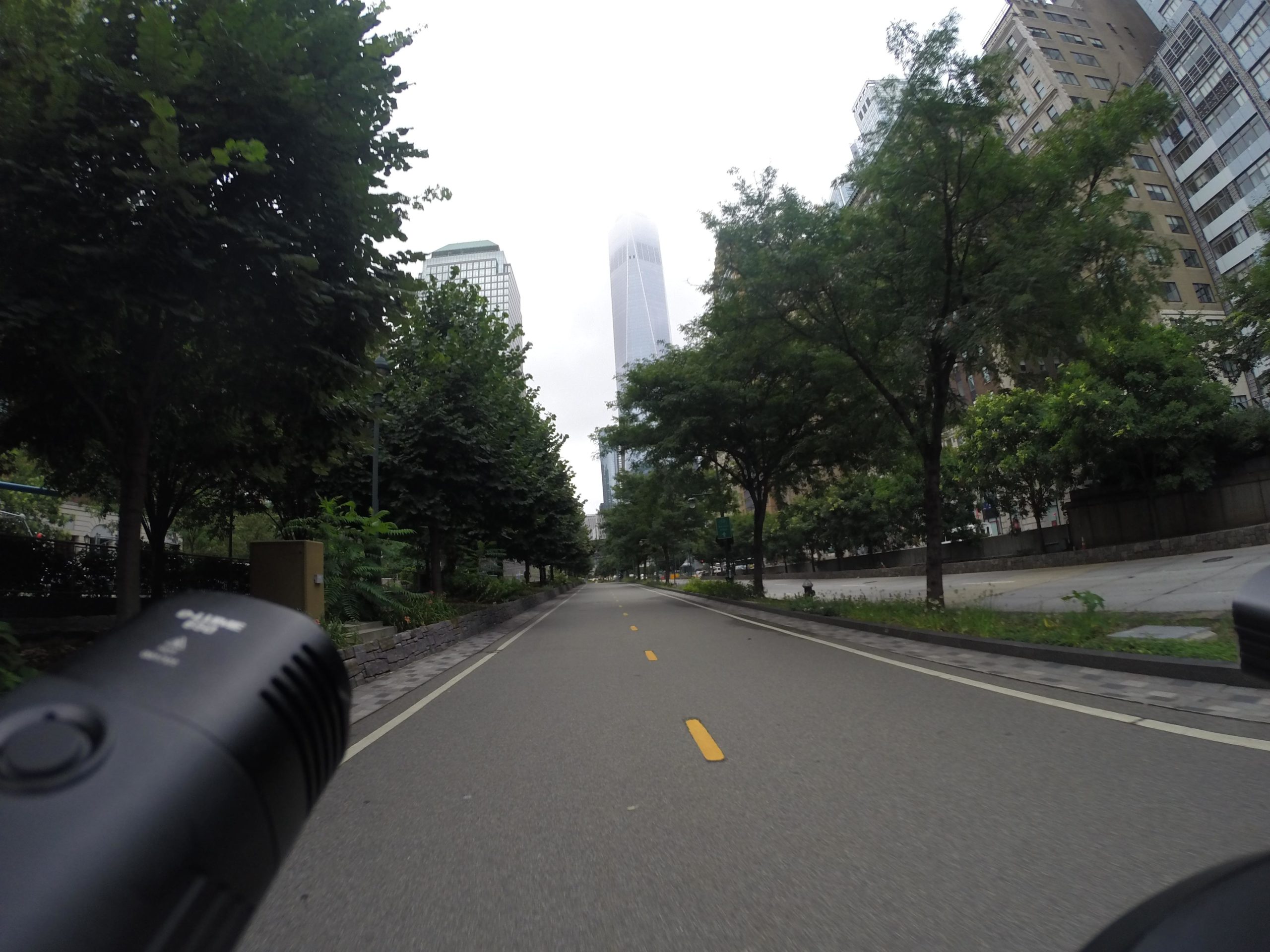 A perspective view of a city street lined with trees, leading towards tall buildings shrouded in mist. The image is taken from a low angle, showcasing the pavement and a portion of a bike handlebar on the left side. The scene conveys a quiet, urban atmosphere with overcast skies above. West Street Greenway mountain bike trail.