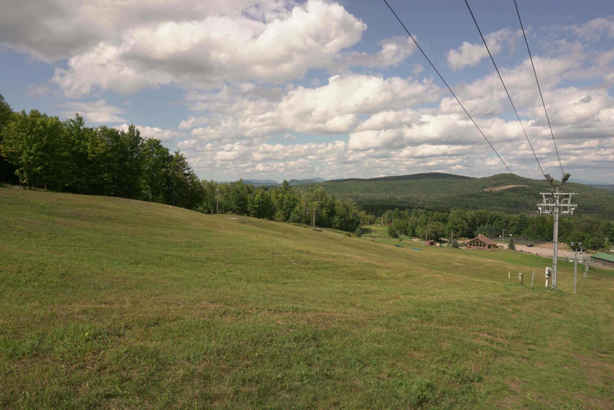 A scenic view of a grassy hillside with trees in the background under a partly cloudy sky. A ski lift tower is visible on the right side of the image, and a building can be seen in the lower part of the frame, surrounded by greenery. Mount Pisgah II: the left side mountain bike trail.