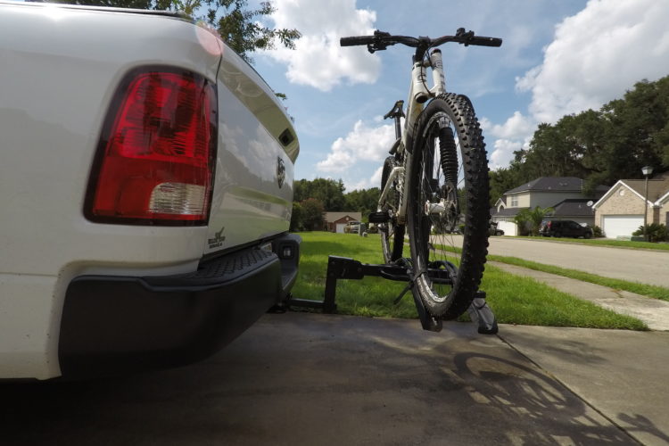 Alt text: A white pickup truck with a mountain bike securely mounted on a bike rack attached to the rear. The background features a grassy area and residential homes under a partly cloudy sky.