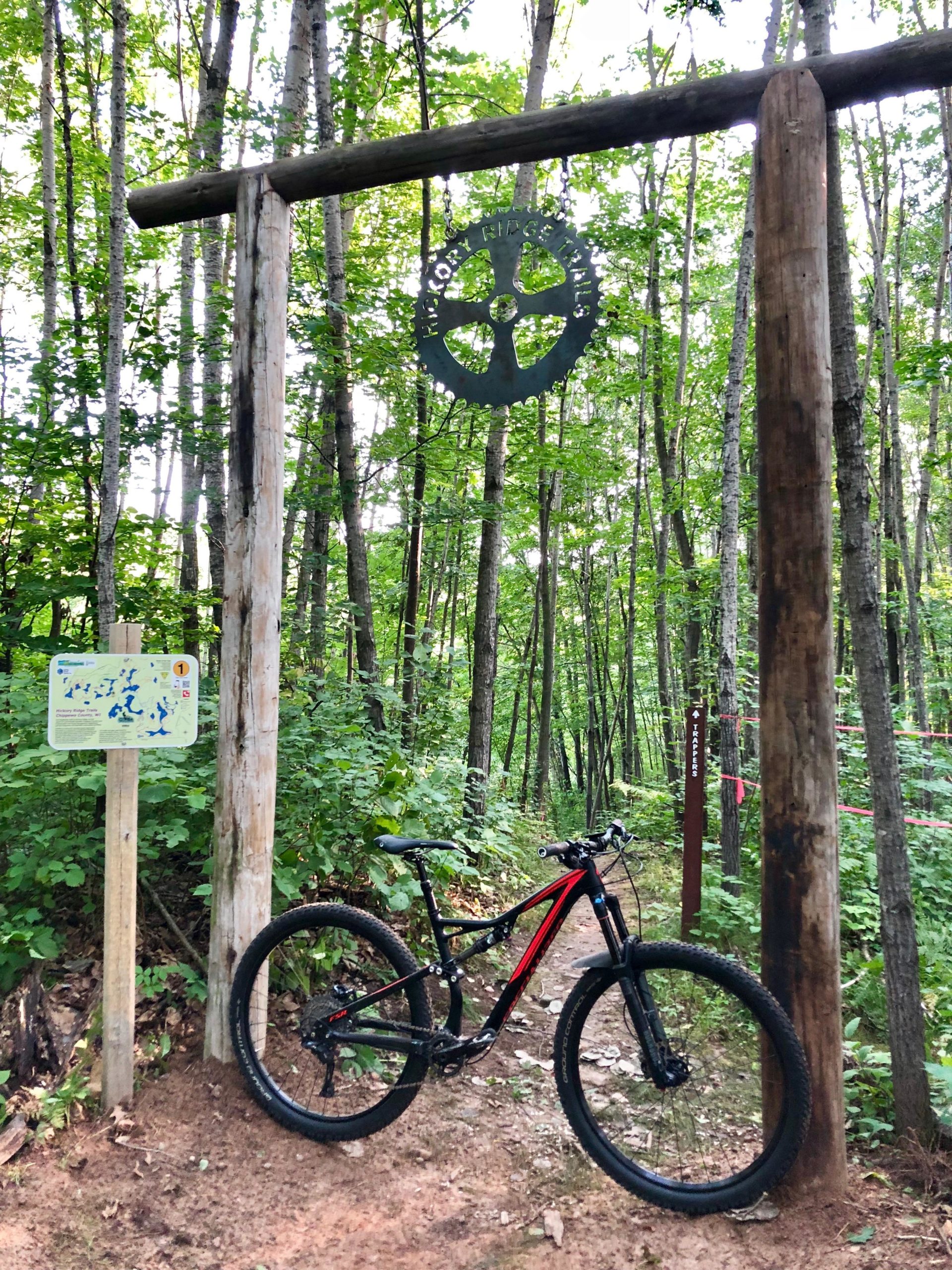 A mountain bike leans against a wooden signpost at the entrance of a forest trail, surrounded by lush green trees. The sign features a map and information about the trail, and a decorative gear-shaped element hangs above, indicating the trail name. The ground is a mix of dirt and rocks, suggesting a natural, rugged environment. Hickory Ridge mountain bike trail.