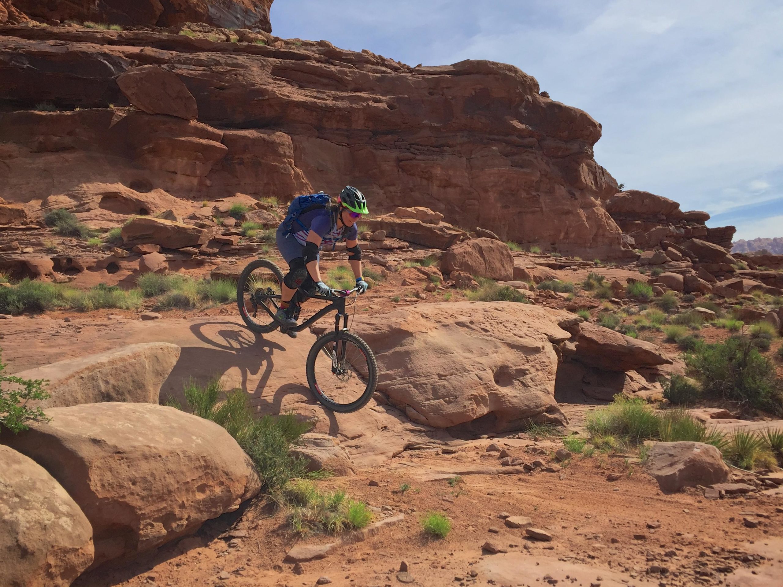 A mountain biker in a colorful outfit and helmet is jumping off a rock formation on a rugged dirt trail, surrounded by red rock terrain and sparse greenery under a clear blue sky. Hymasa mountain bike trail.