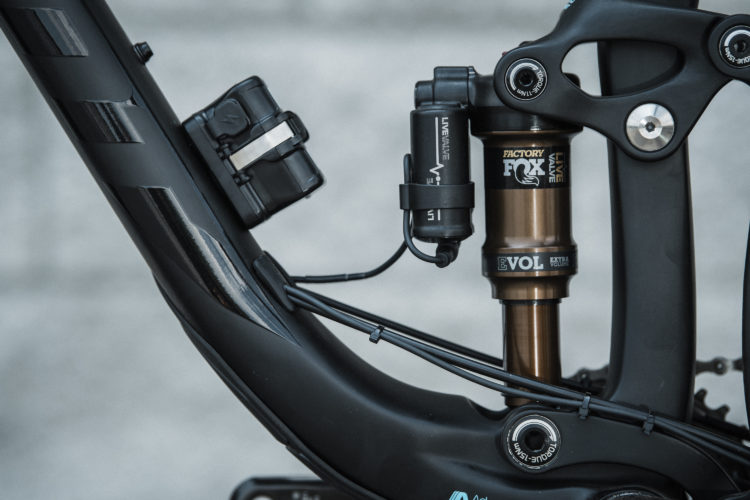 Close-up view of a bicycle's suspension system, featuring a black frame with shiny accents, a brown shock absorber labeled "FOX," and various components such as a remote system labeled "LIVE VALVE." The image showcases the intricate details of the bike's design and engineering.
