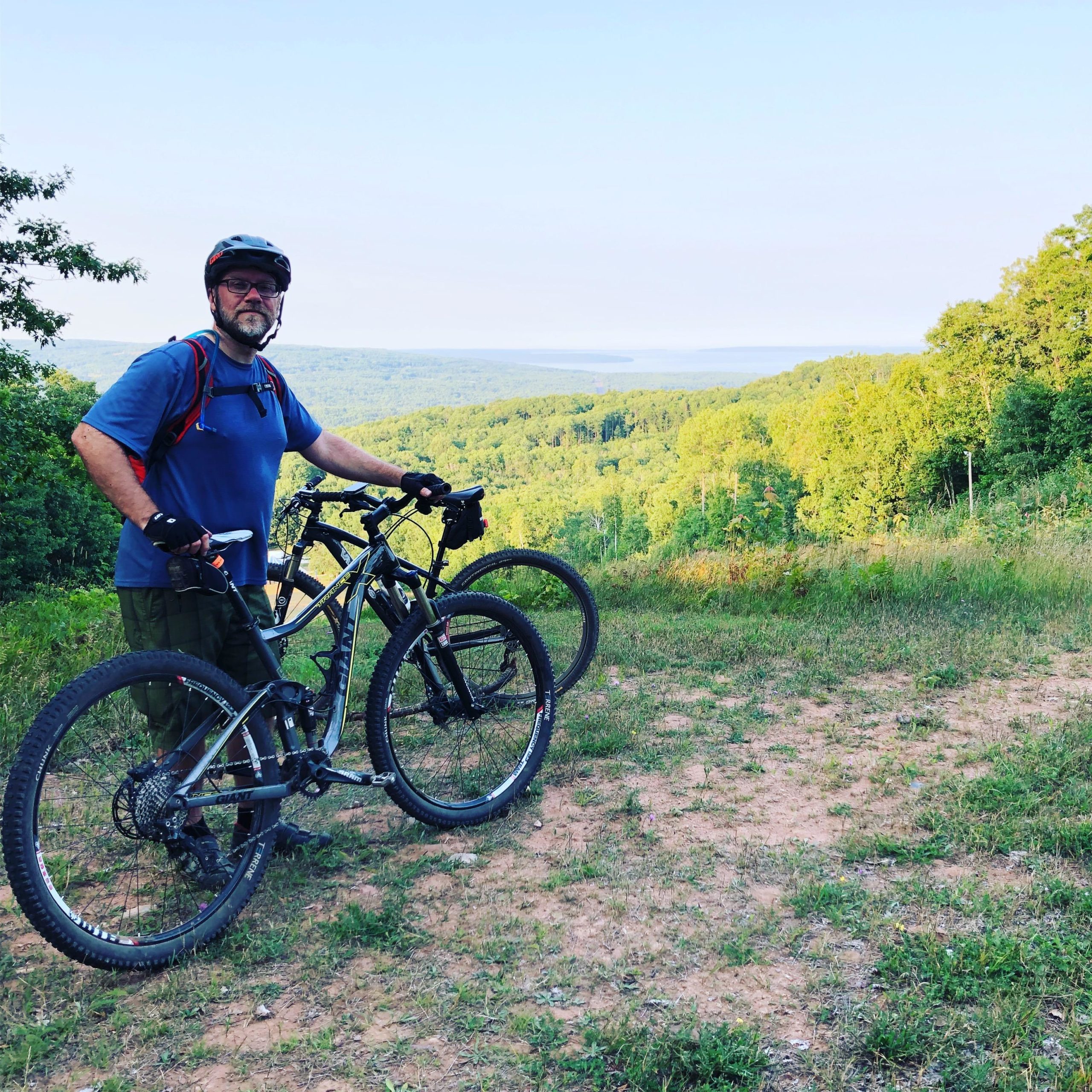 A person in a blue shirt and helmet stands beside two mountain bikes on a grassy hillside, overlooking a lush green landscape with rolling hills and a distant lake under a clear sky. Mt Ashwabay Singletrack mountain bike trail.