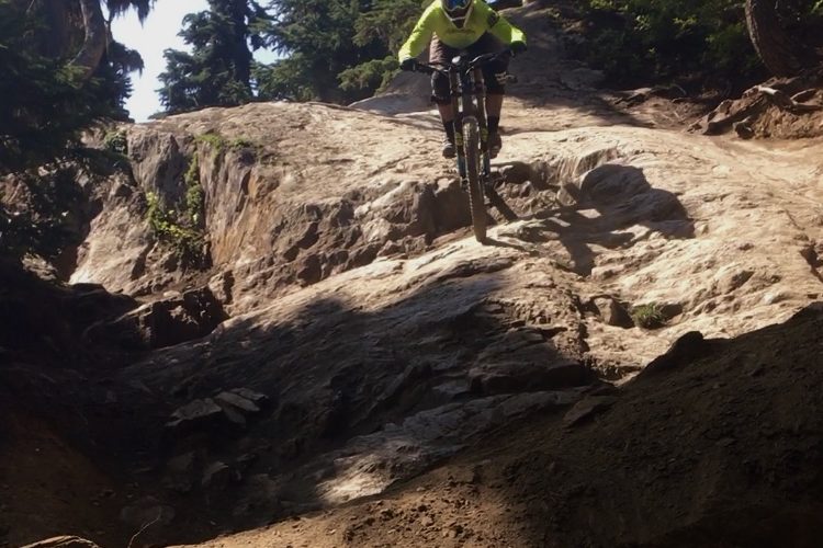 A mountain biker in a bright yellow long-sleeve jersey navigates a rocky terrain with trees in the background. The biker is airborne, showcasing skill as they descend a slope.