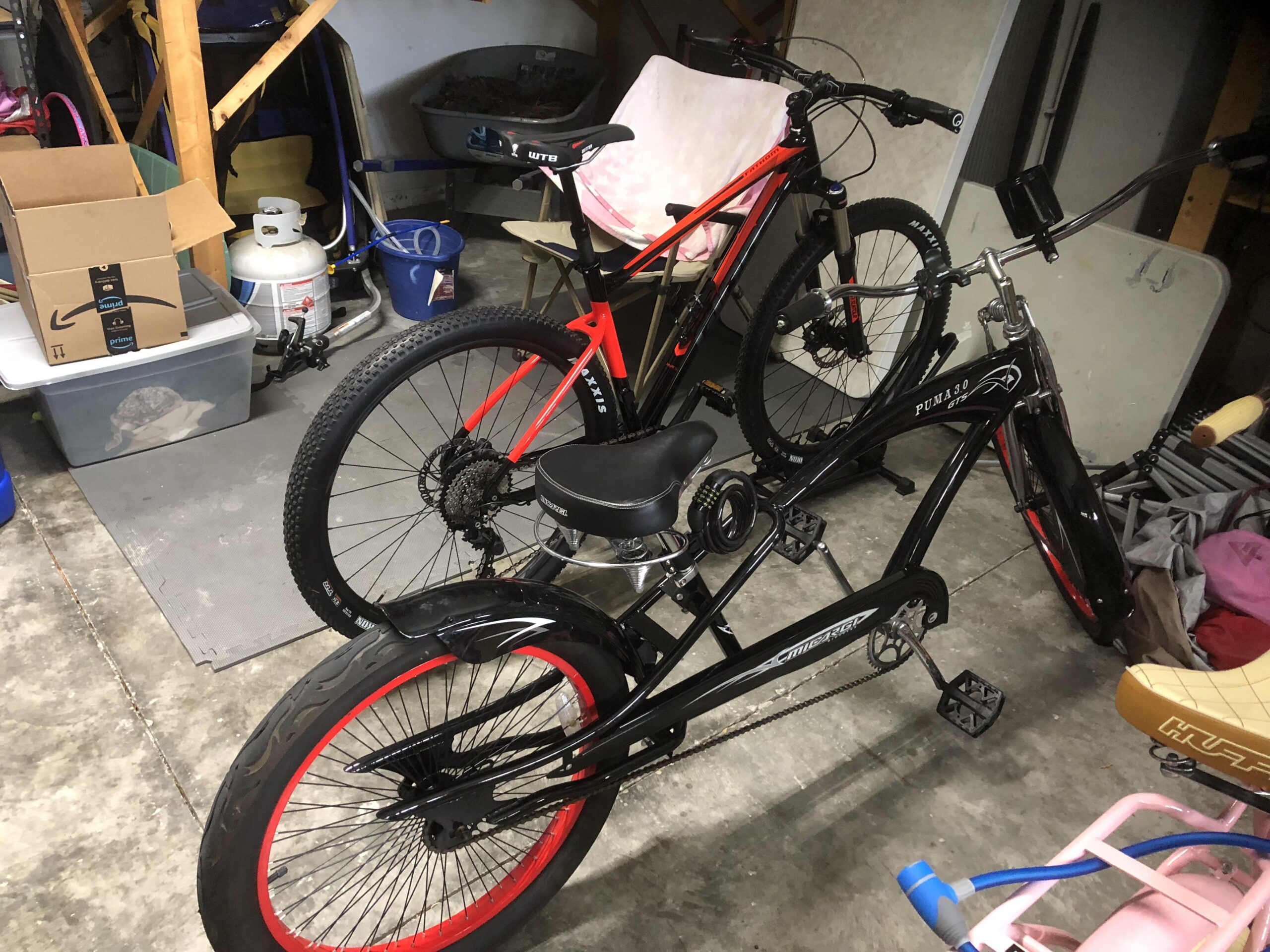 Giant Giant Fathom 2 29er: Two bicycles are parked inside a garage. One bike is a black cruiser with red accents, featuring a wide rear wheel. The other bike is a red mountain bike with thick tires. In the background, there are cardboard boxes, a propane tank, and various storage containers, along with some clutter on the floor. The garage walls and floor are concrete, and the space appears functional but somewhat disorganized.