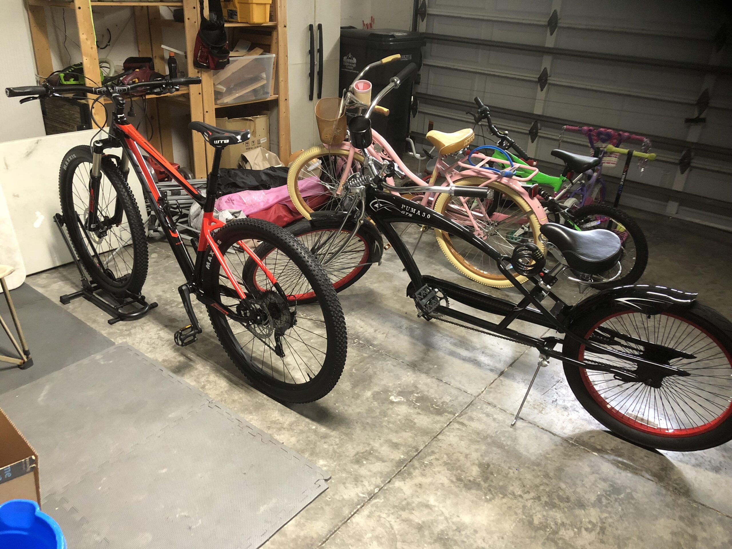 Giant Giant Fathom 2 29er: A garage interior featuring two bikes: a black and red mountain bike with thick tires and a sleek design, and a black cruiser bike with a low frame and stylish curved handlebars. In the background, there are several colorful children's bikes, including pink and green models, and various items stored on shelves. The floor is concrete with a puzzle mat section.